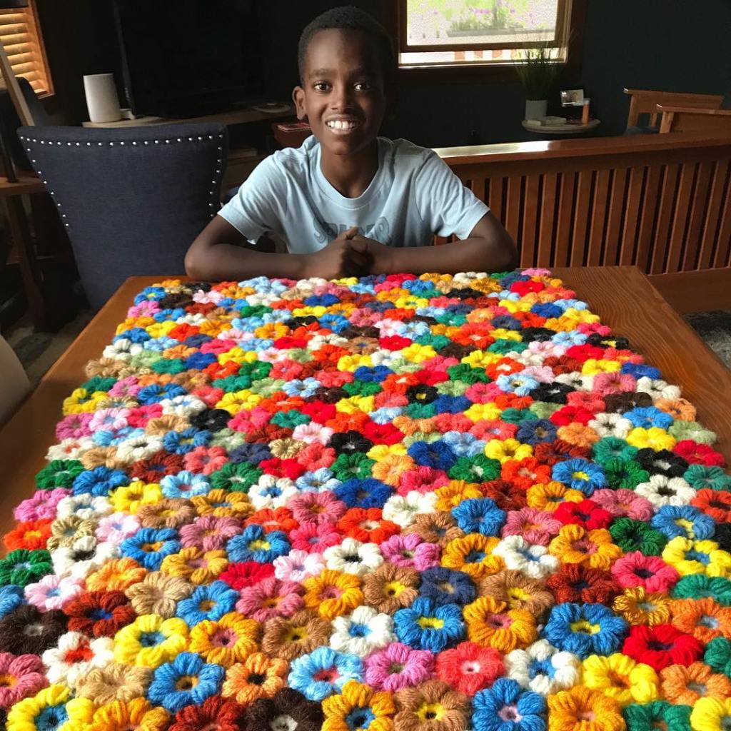 Boy sitting at a table with a large crocheted blanket of multicolored flowers.