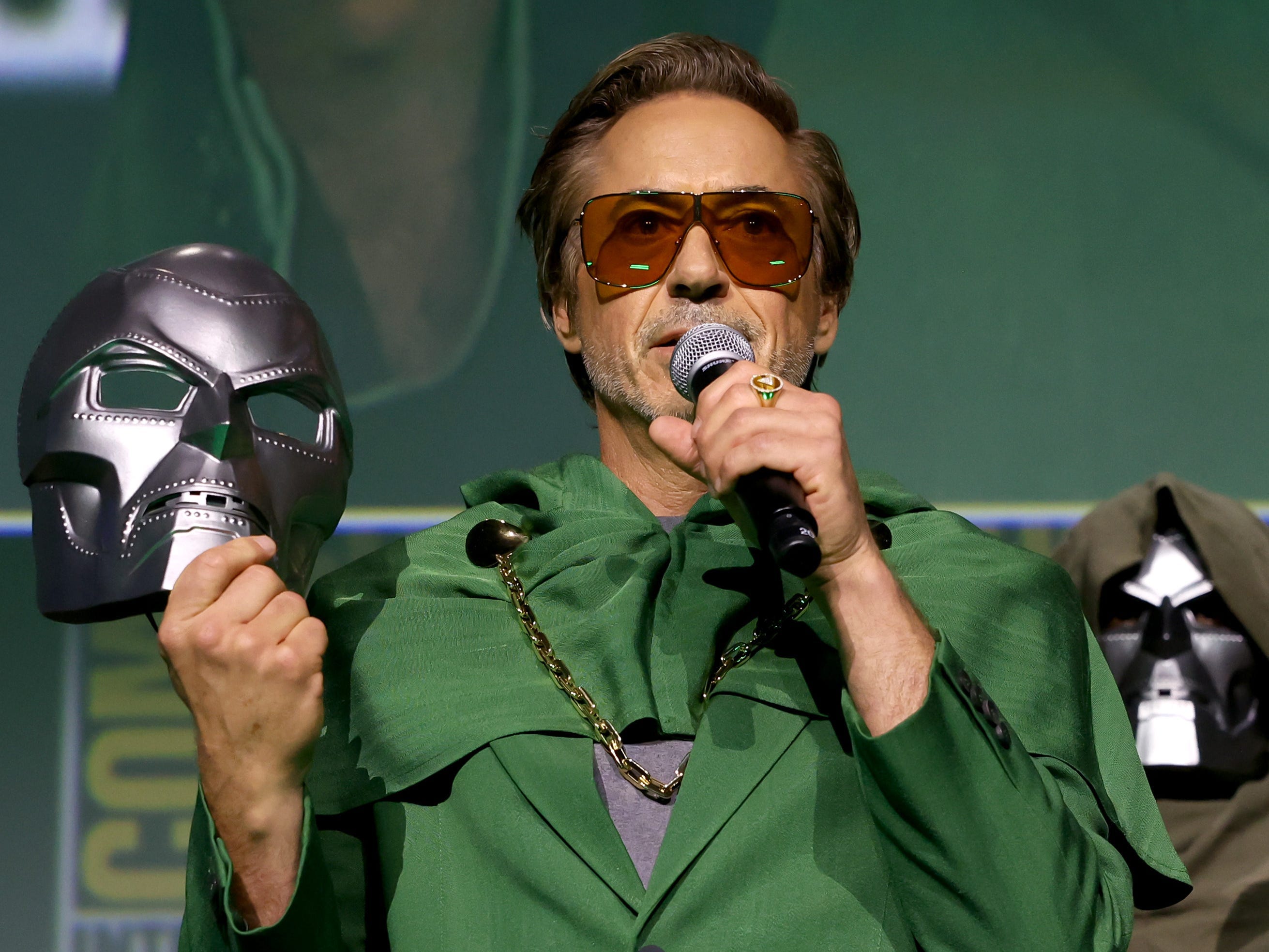 Robert Downey Jr. in a green suit holding a silver mask and a microphone.