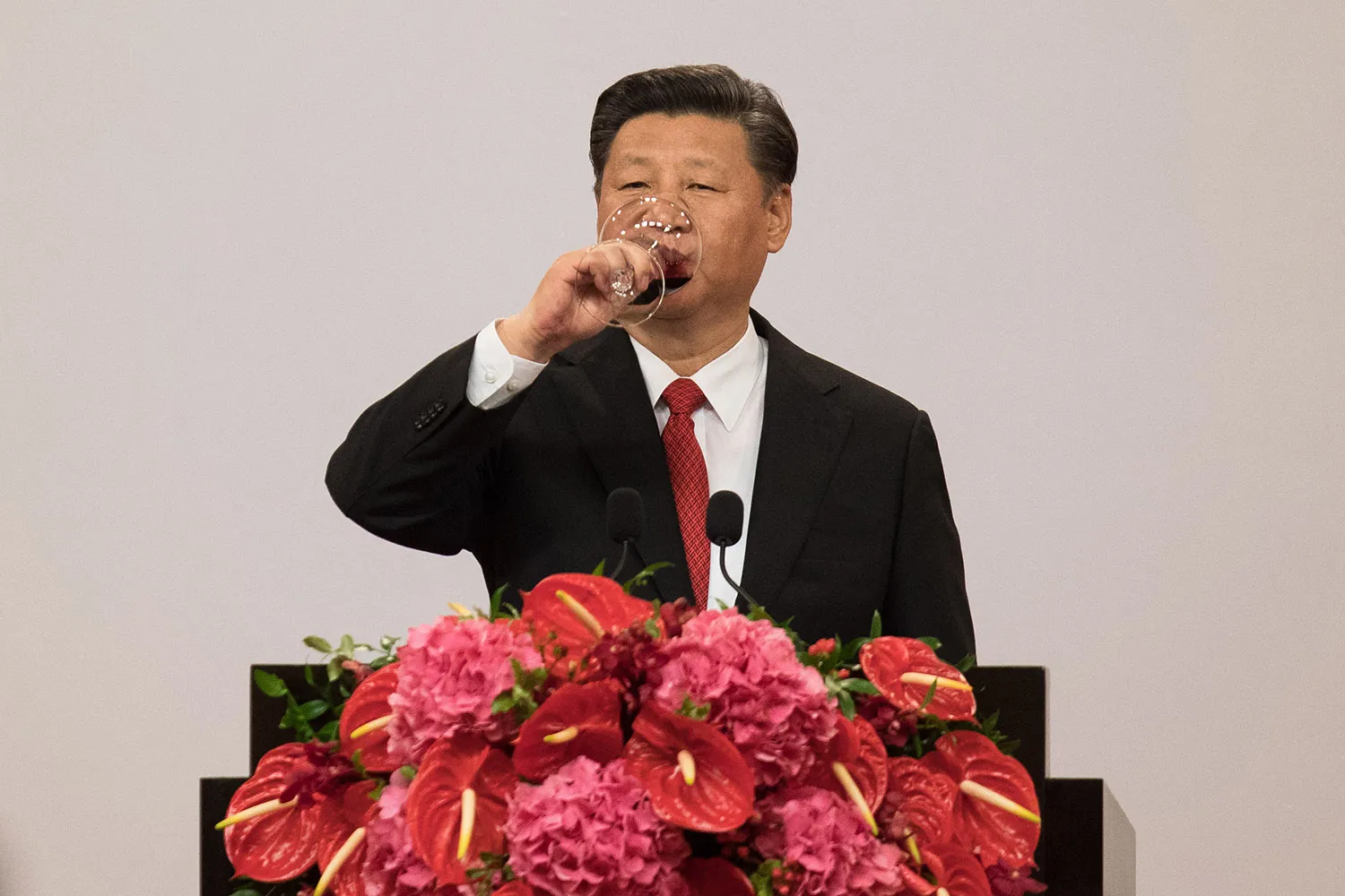 Xi Jinping in a suit drinking from a wine glass behind a flower-covered lectern.