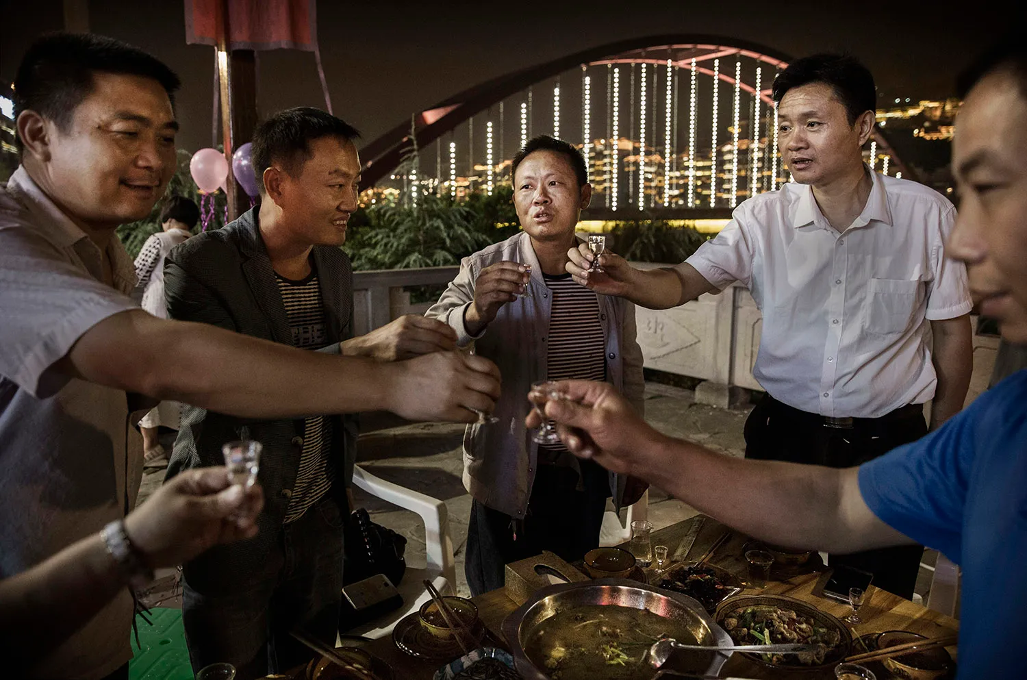 Men raises small glasses in a toast as they gather around a table.