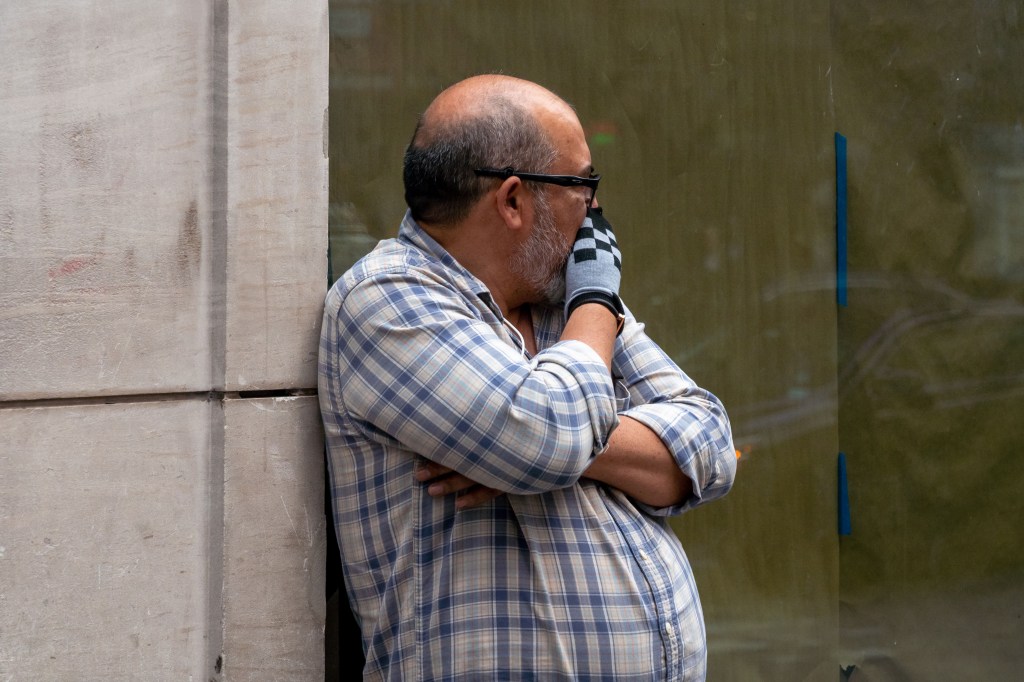 Man in plaid shirt standing outdoors, covering his mouth with a glove due to smoke.
