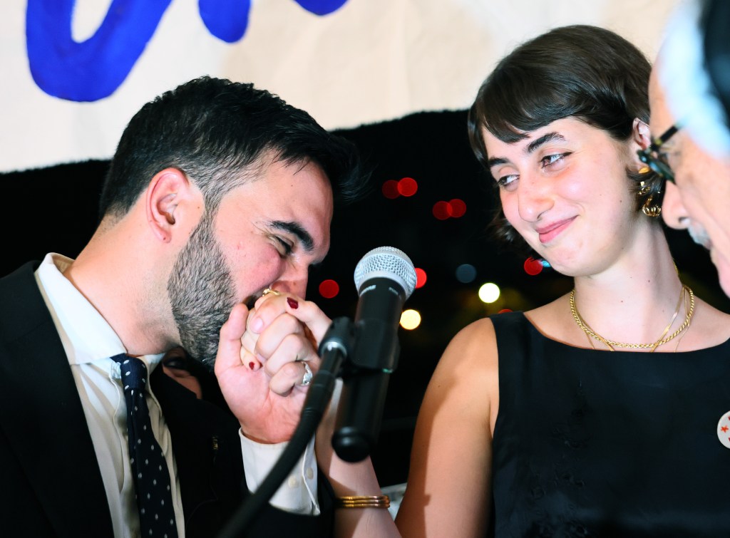 mamdani kisses his wife's hand while they stand at a micro phone