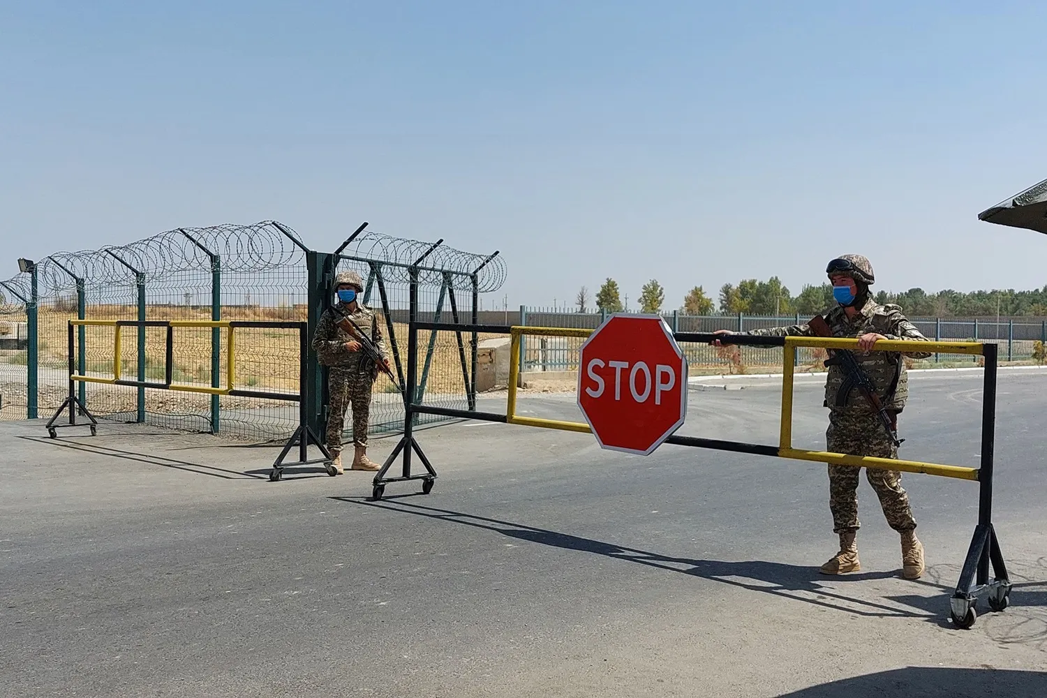 Two Uzbek soldiers, both wearing camouflage and helmets and holding rifles, stand on either side of a metal gate with a stop sign at its center. Behind the fence is a flat field, and farther in the distance are trees and a blue sky.