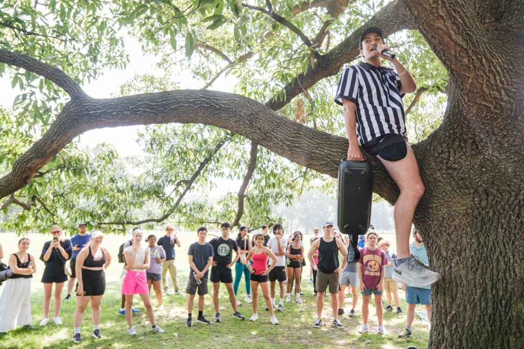 A man in a referee shirt sits in a tree speaking into a microphone to a group of people.