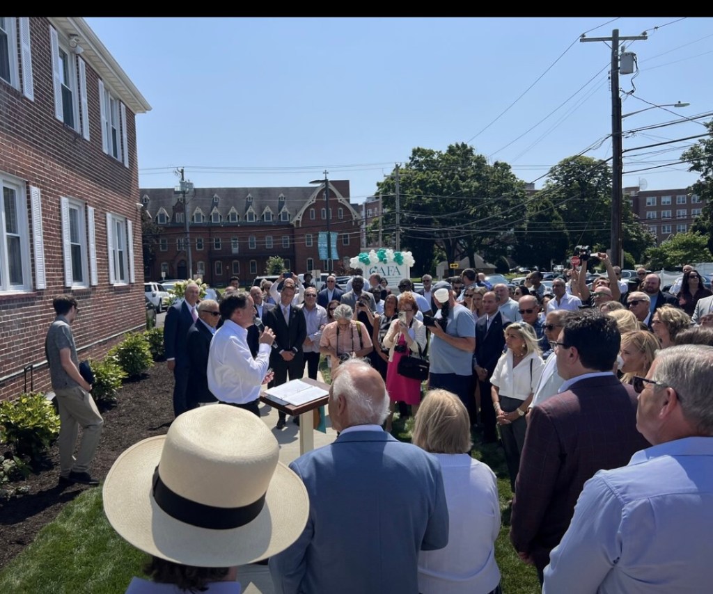 Large group of people gathered outdoors for an event, a man speaking at a podium.