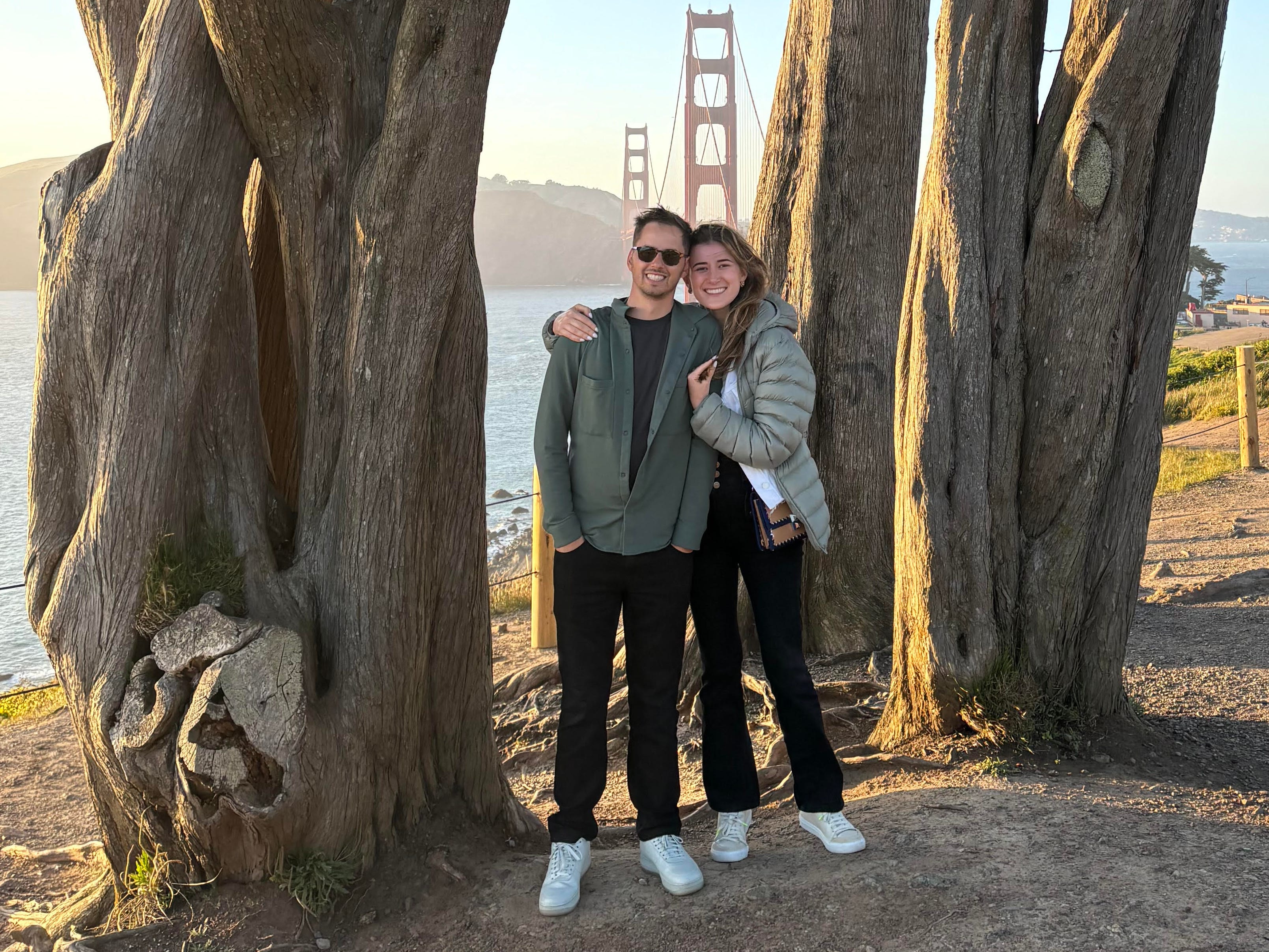 a man and woman standing in between two large trees with San Francisco's Golden Gate Bridge in the background