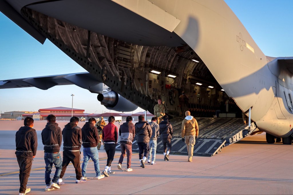 Detainees in chains being escorted toward a military transport plane.