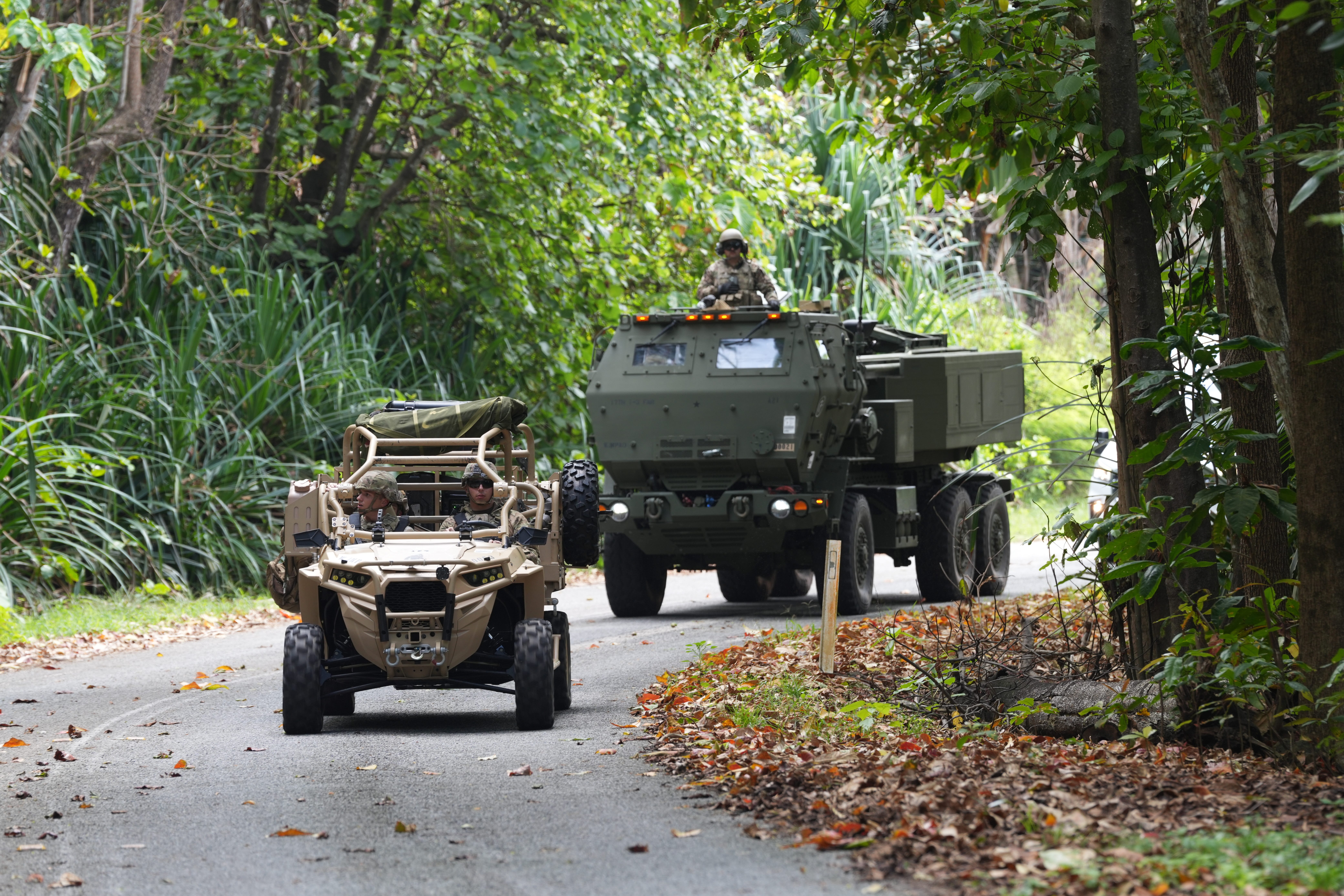 A green truck drives down a jungle road behind a beige vehicle.