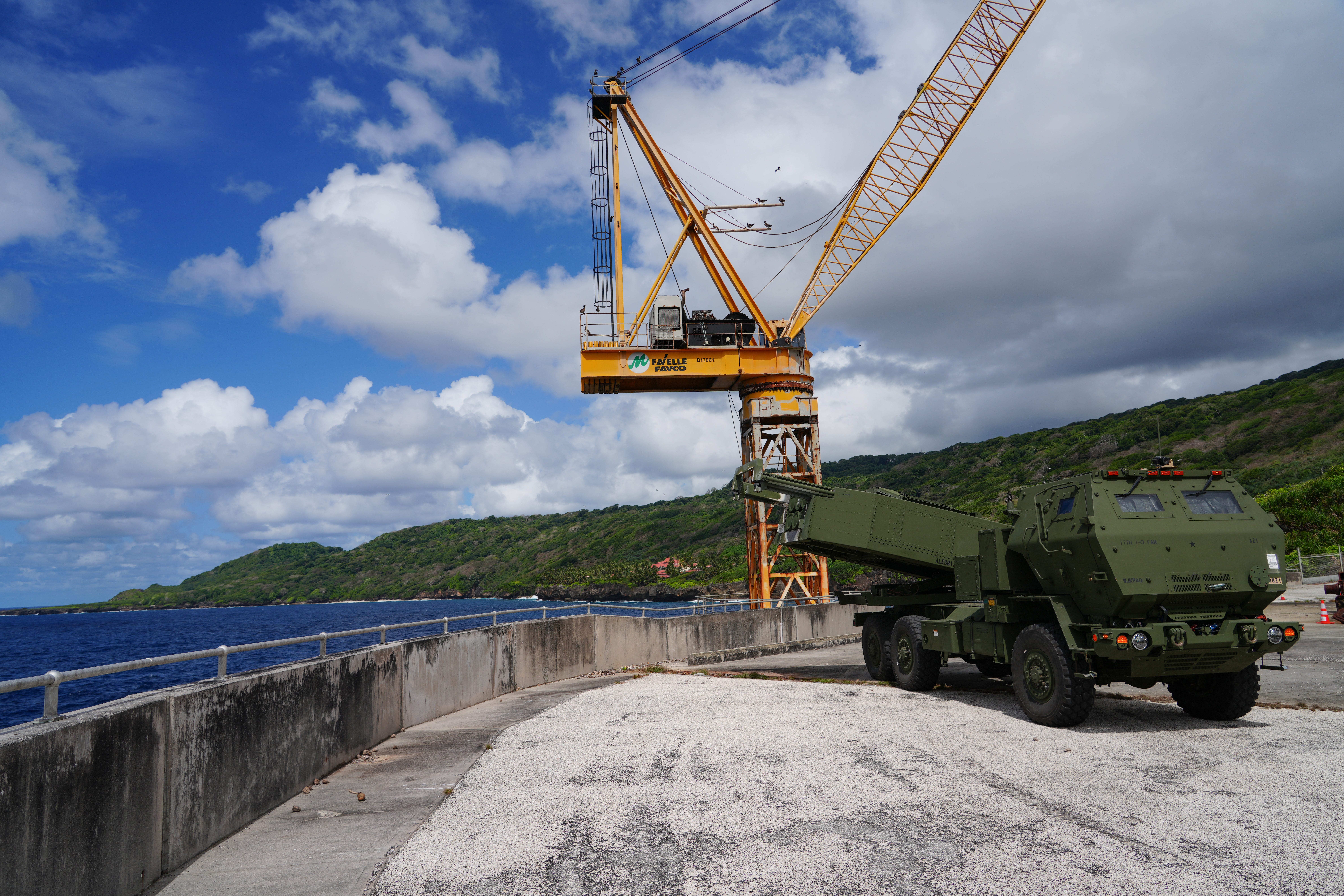 A green truck sits with a missile launcher pointed left on top of it. Behind the truck is a yellow crane. The truck is overlooking blue ocean water. A green mountainous landscape is visible in the background.
