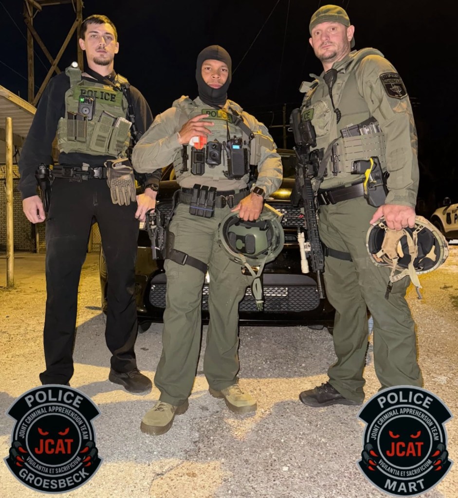 Three law enforcement officers in tactical gear stand in front of a police vehicle.