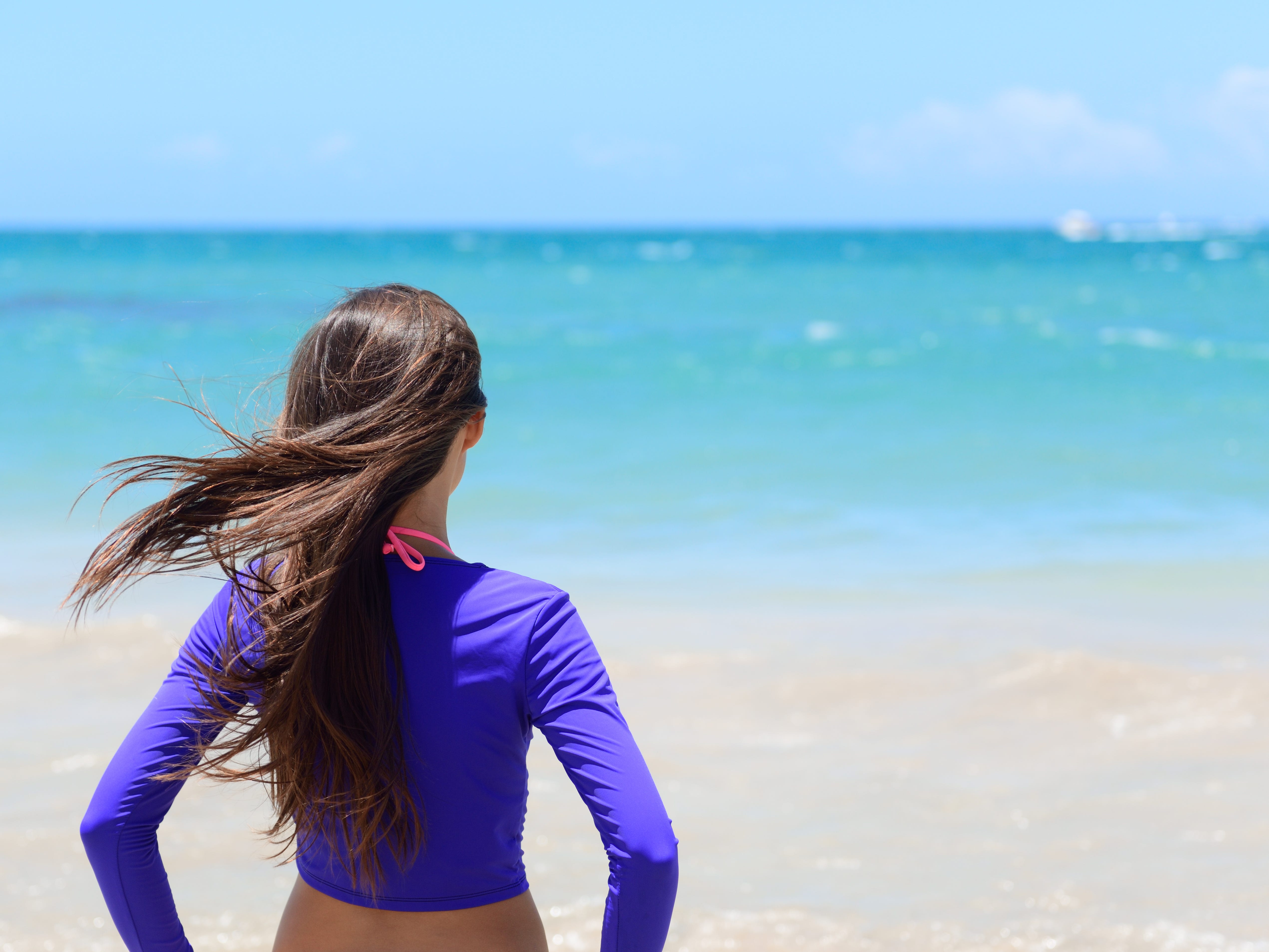 A woman standing with her back to the camera looking out into the ocean wearing a purple water-shirt.