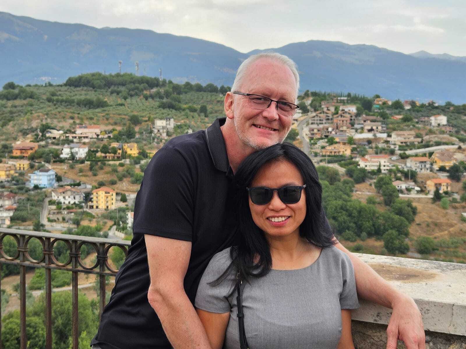 Todd Kleperis and his wife embracing in front of a hilly scene in Italy.