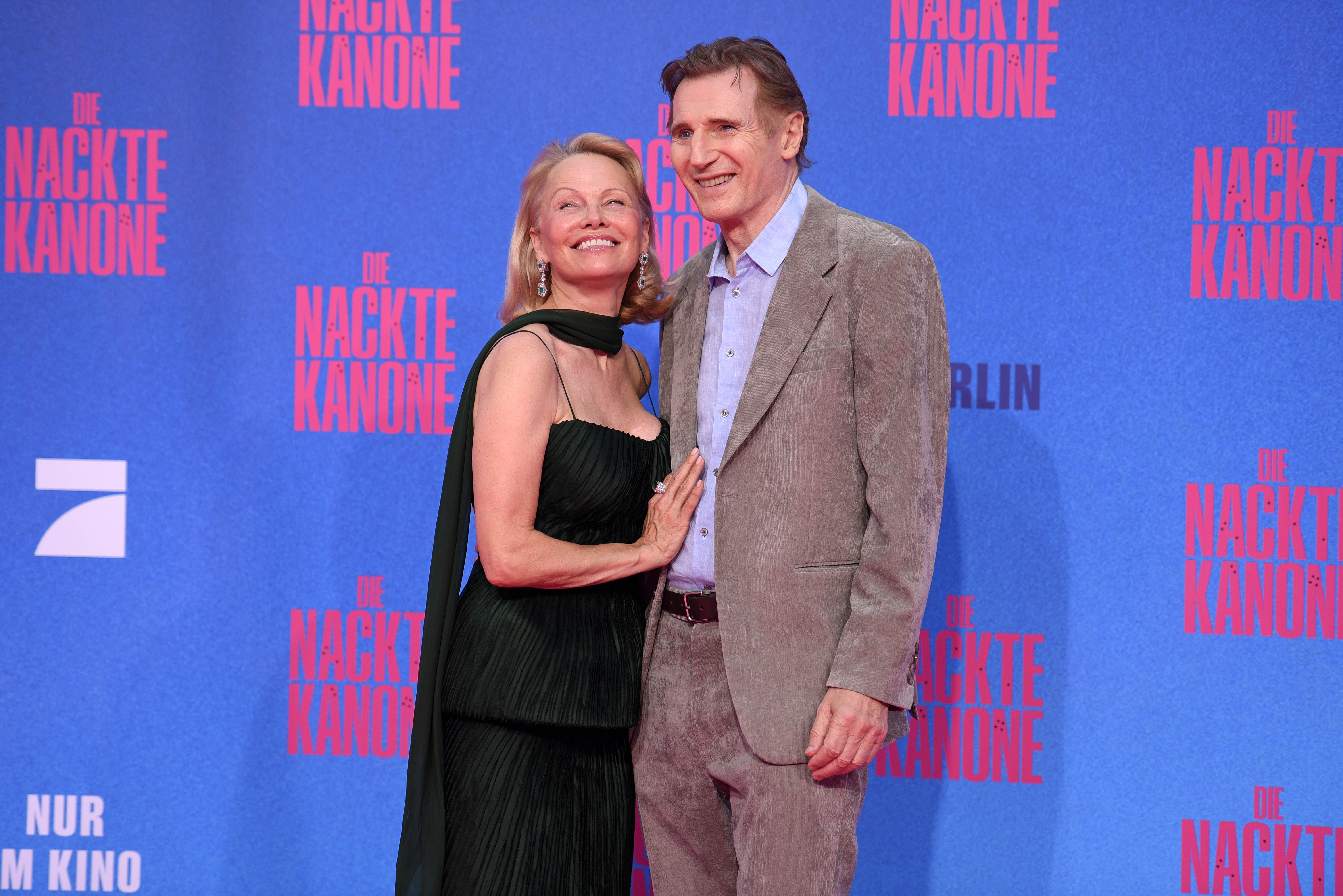 Pamela Anderson in a black dress standing next to Liam Neeson in a grey suit