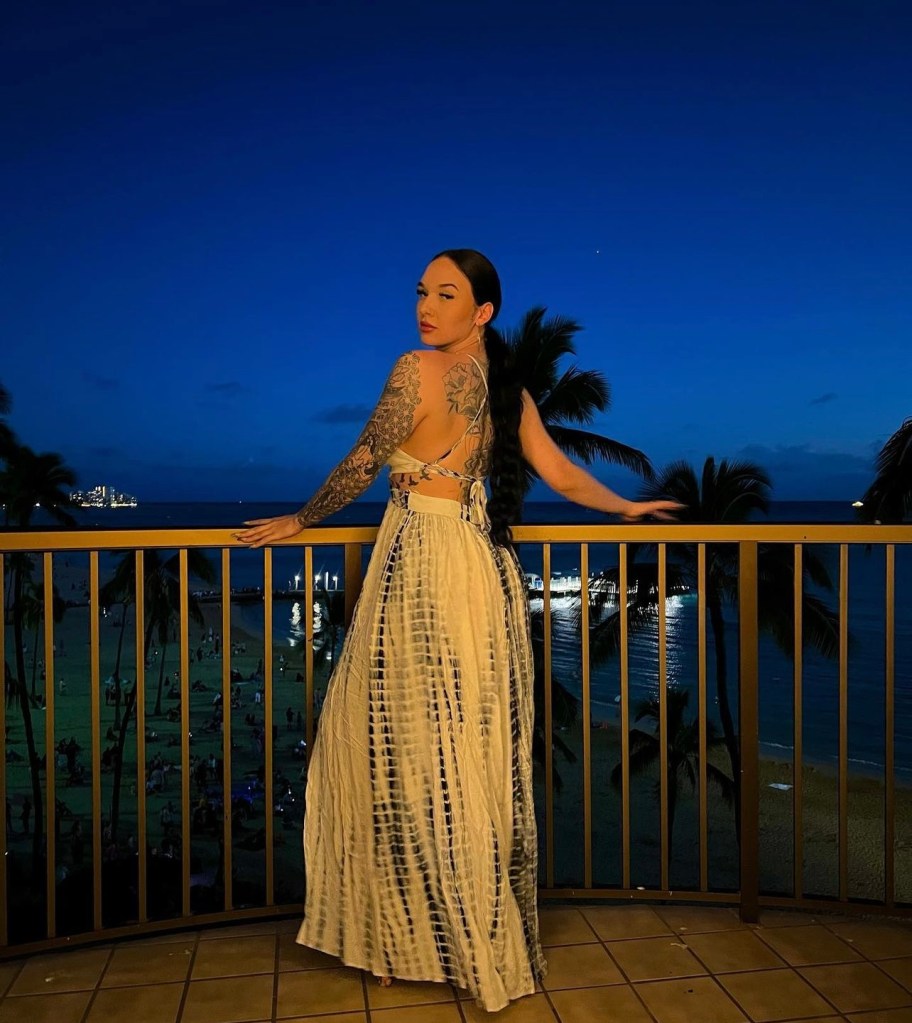Woman in tie-dye dress on balcony overlooking ocean at night.