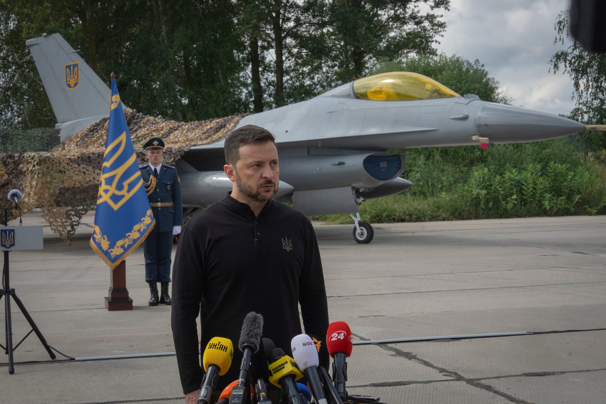 Ukrainian President Volodymyr Zelenskyy stands outside in front of microphones with a Ukrainian Air Force F-16 fighter jet behind him.