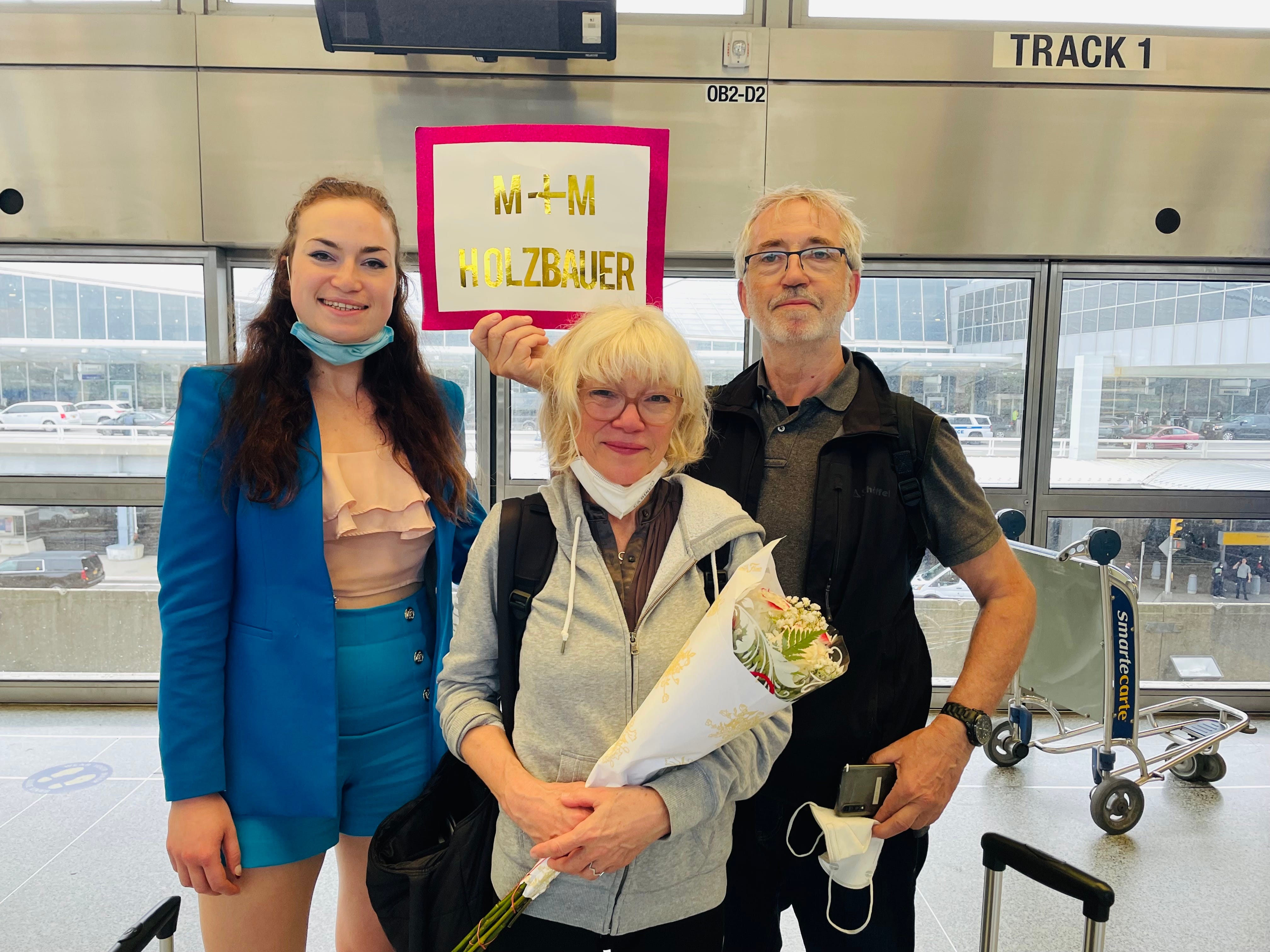 The author with her parents at the airport, they are holding flowers and a sign.