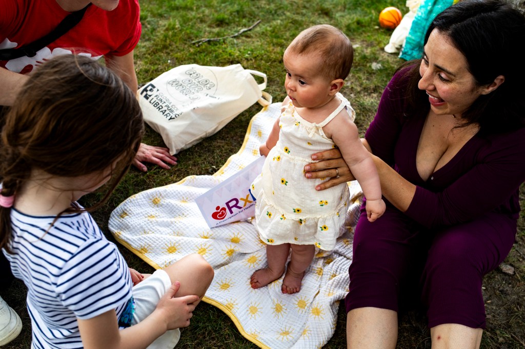 A mother holds her baby daughter while sitting on a blanket on the grass.  A young girl sits nearby.