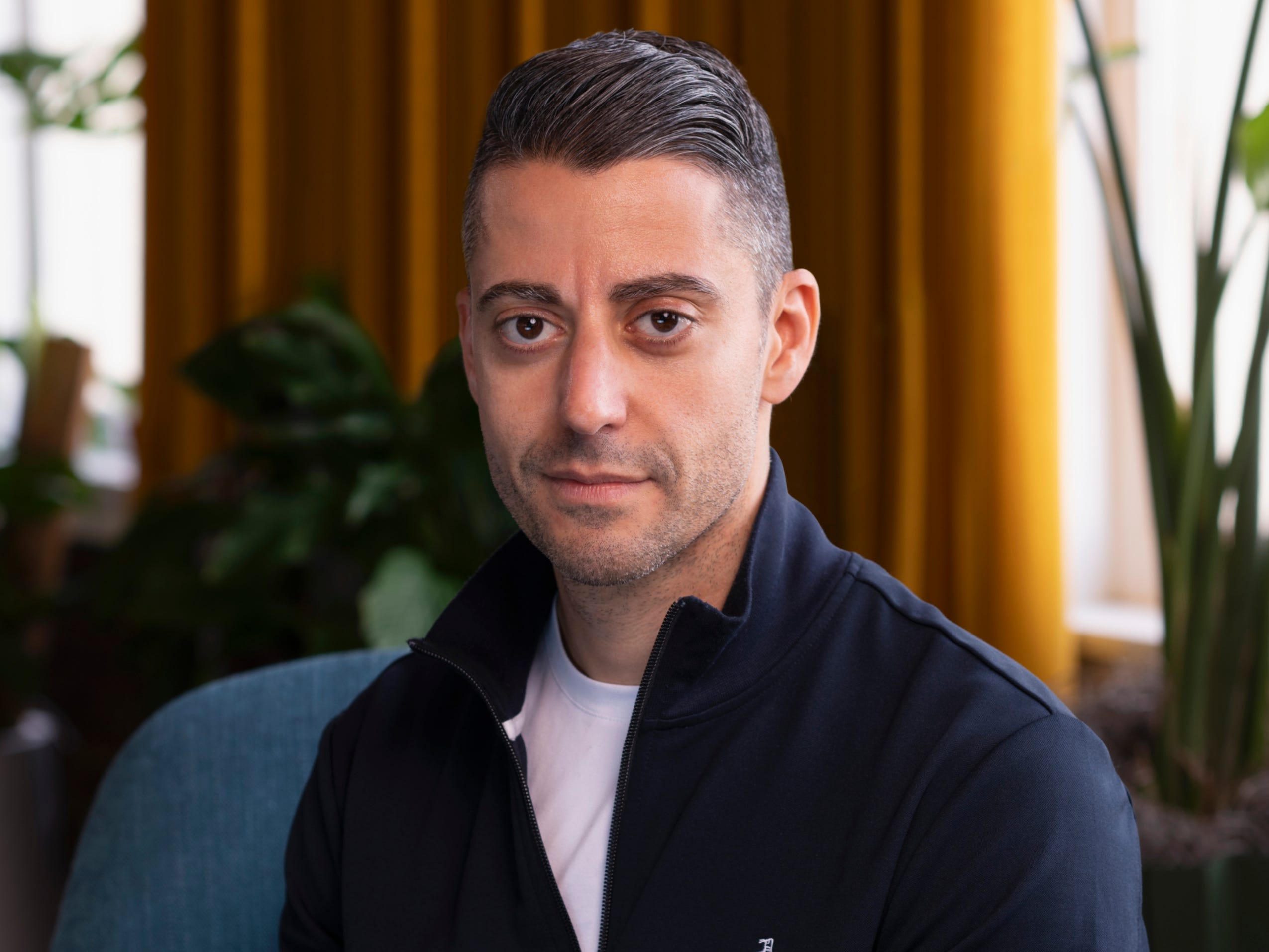 Shaunt Voskanian, wearing a zip-up shirt and sitting in a blue chair in front of plants.