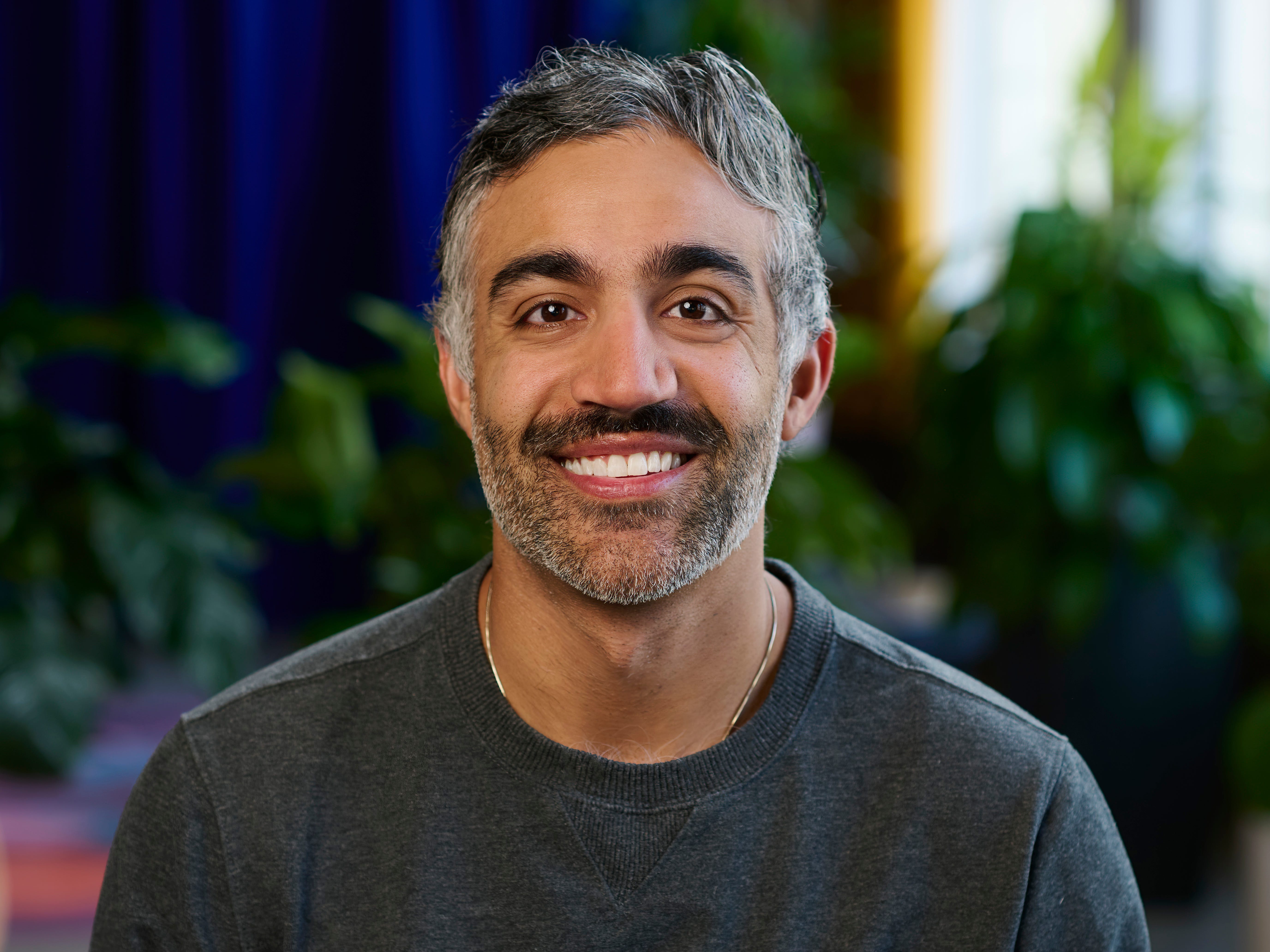 Praveer Melwani, wearing a grey shirt, silver necklace, and seated in front of some plants.