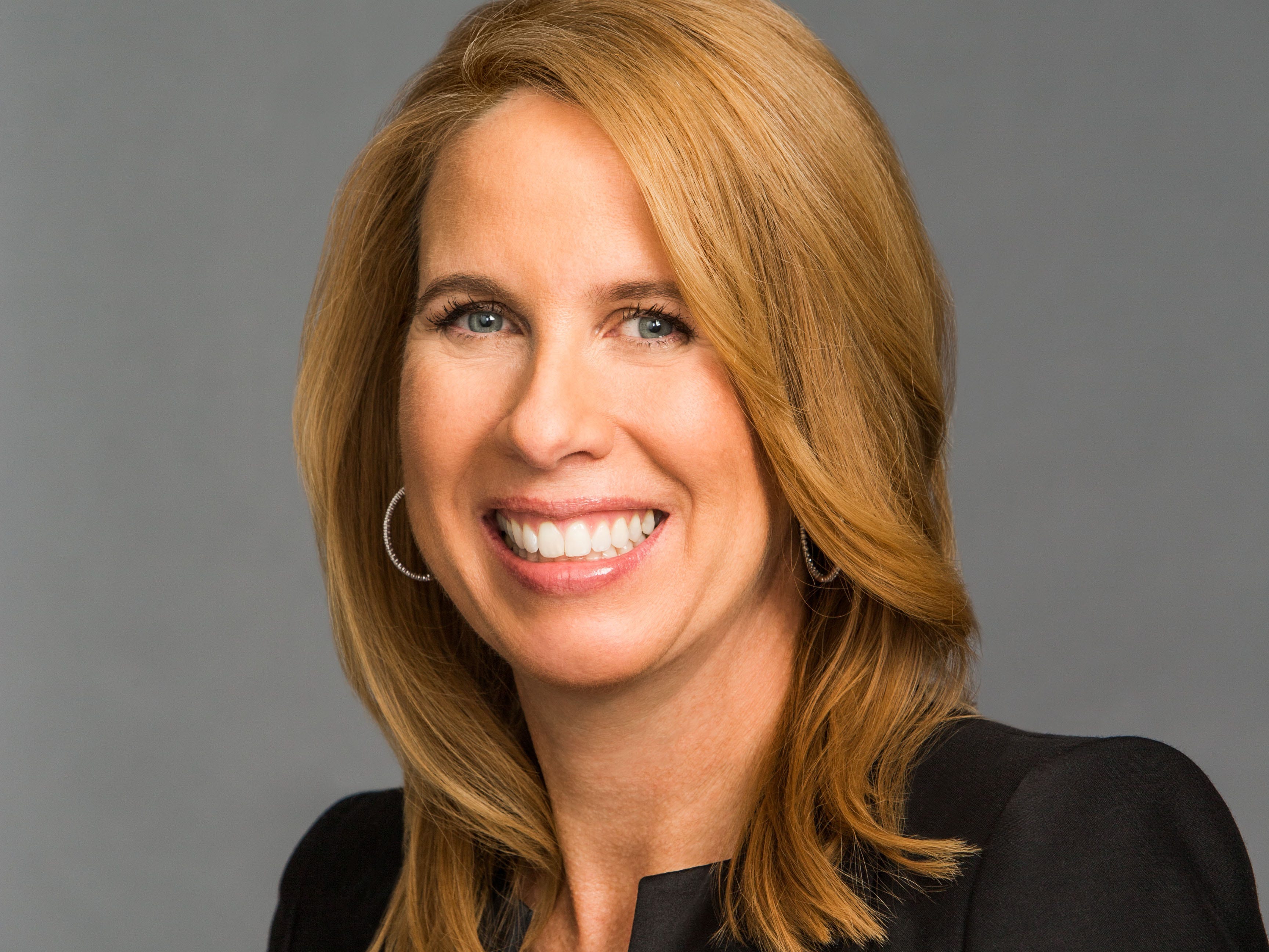 Lynn Vojvodich, wearing a black blazer and white shirt in front of a grey background.