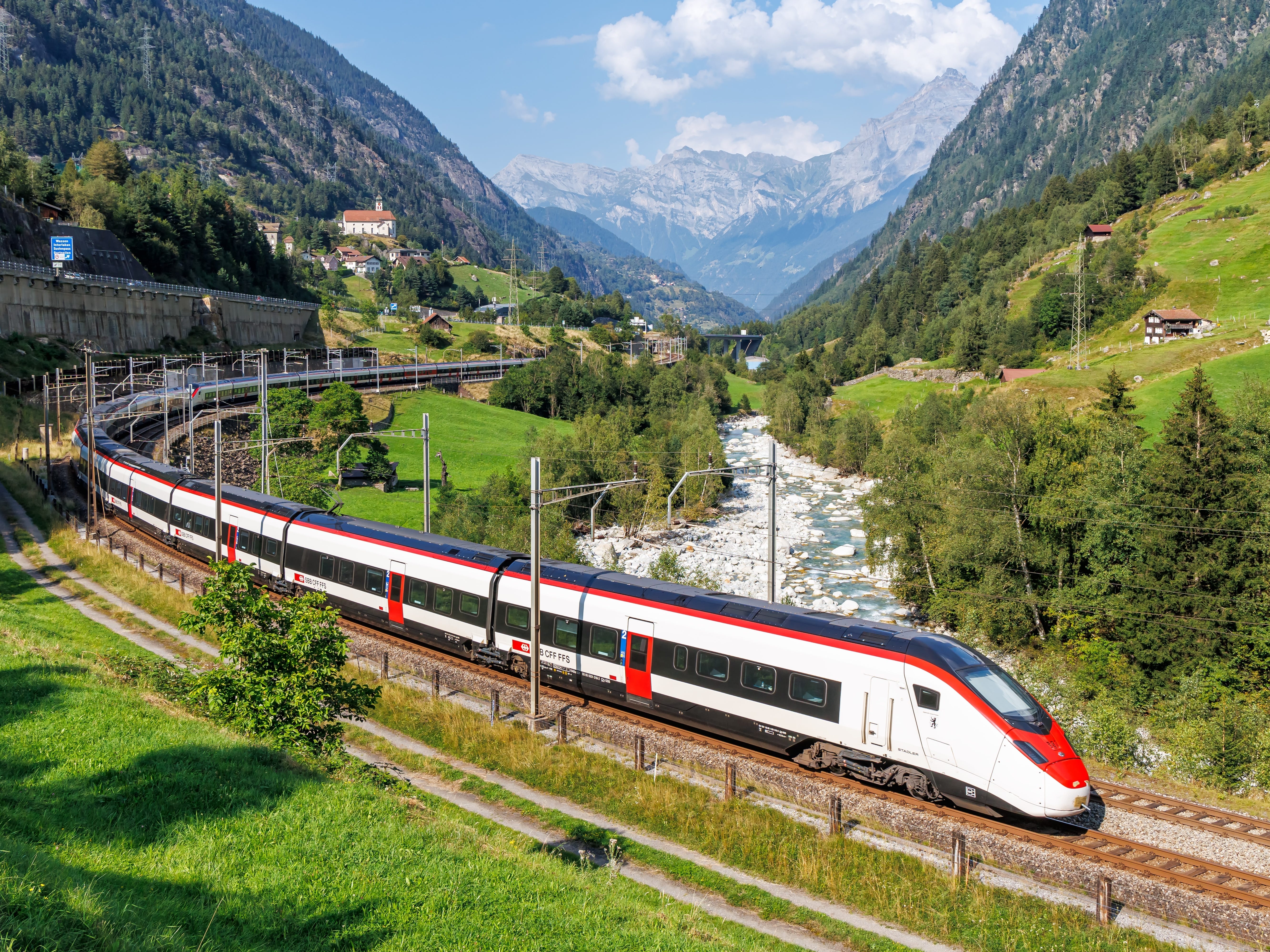 A Gotthard passenger train on route through the Swiss Alps in Wassen, Switzerland. There are trees, mountains, hills, and a stream.