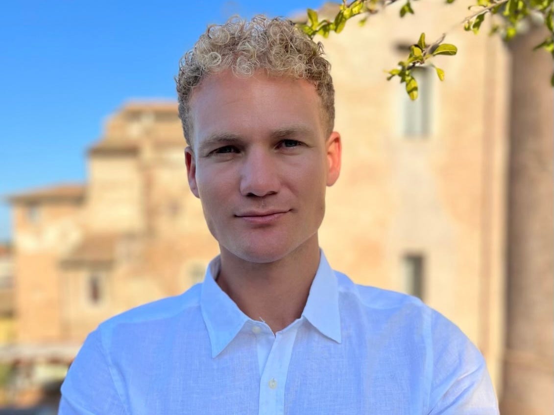 A picture of Sam Awrabi, standing in front of old buildings and wearing a white oxford shirt with his arms crossed.