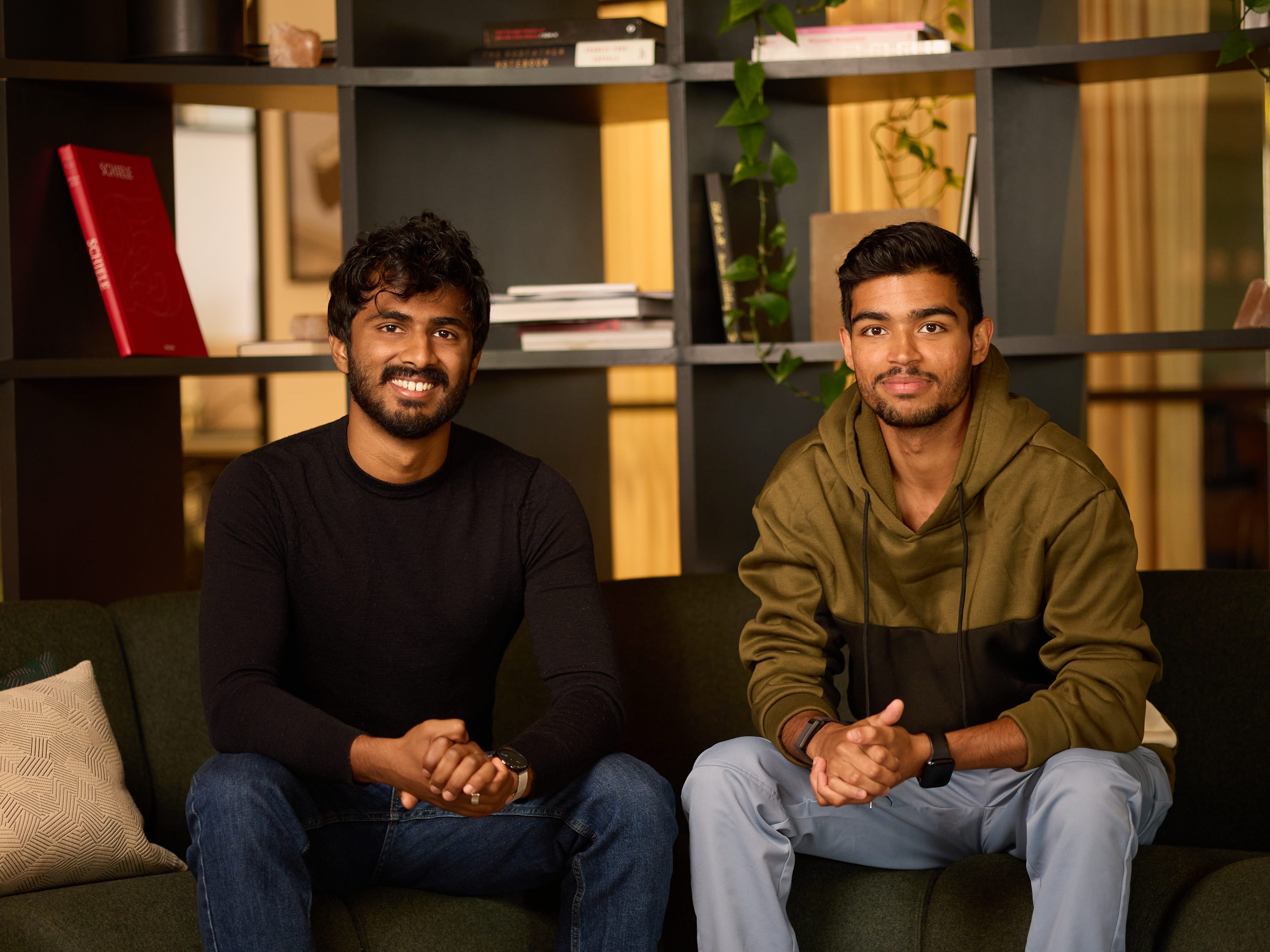 Roop Pal, in a black sweater, and Puneet Sukhija, in a green hoodie, sitting on a sofa smiling with their hands clasped in their laps.