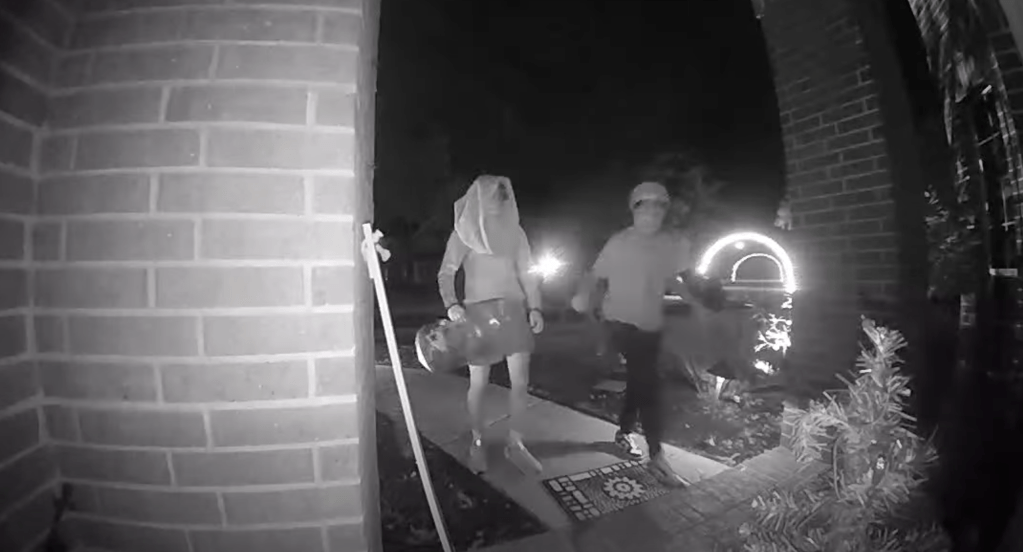 Nighttime security camera footage of two individuals carrying a skateboard and a bag walking away from a house.