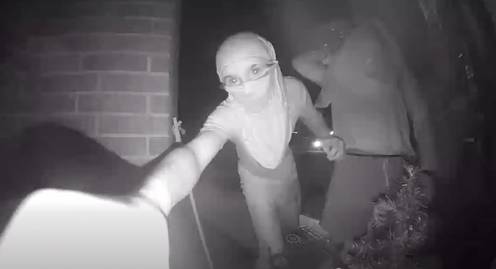 Night vision security camera footage of two masked individuals near a doorway.