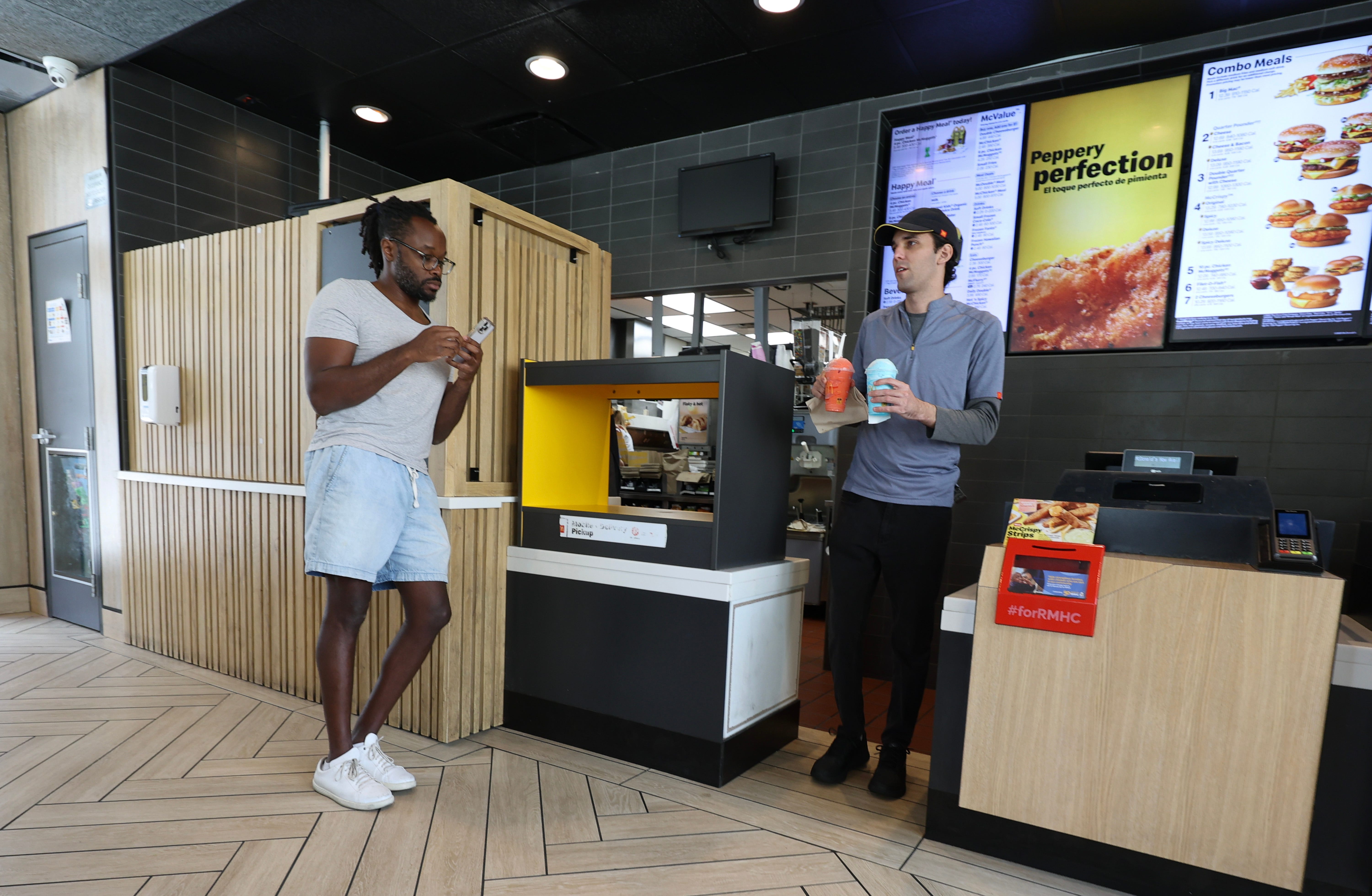 A McDonalds employee brings two beverages out to a customer as a customer waits and looks at his phone while standing next to the pickup area.