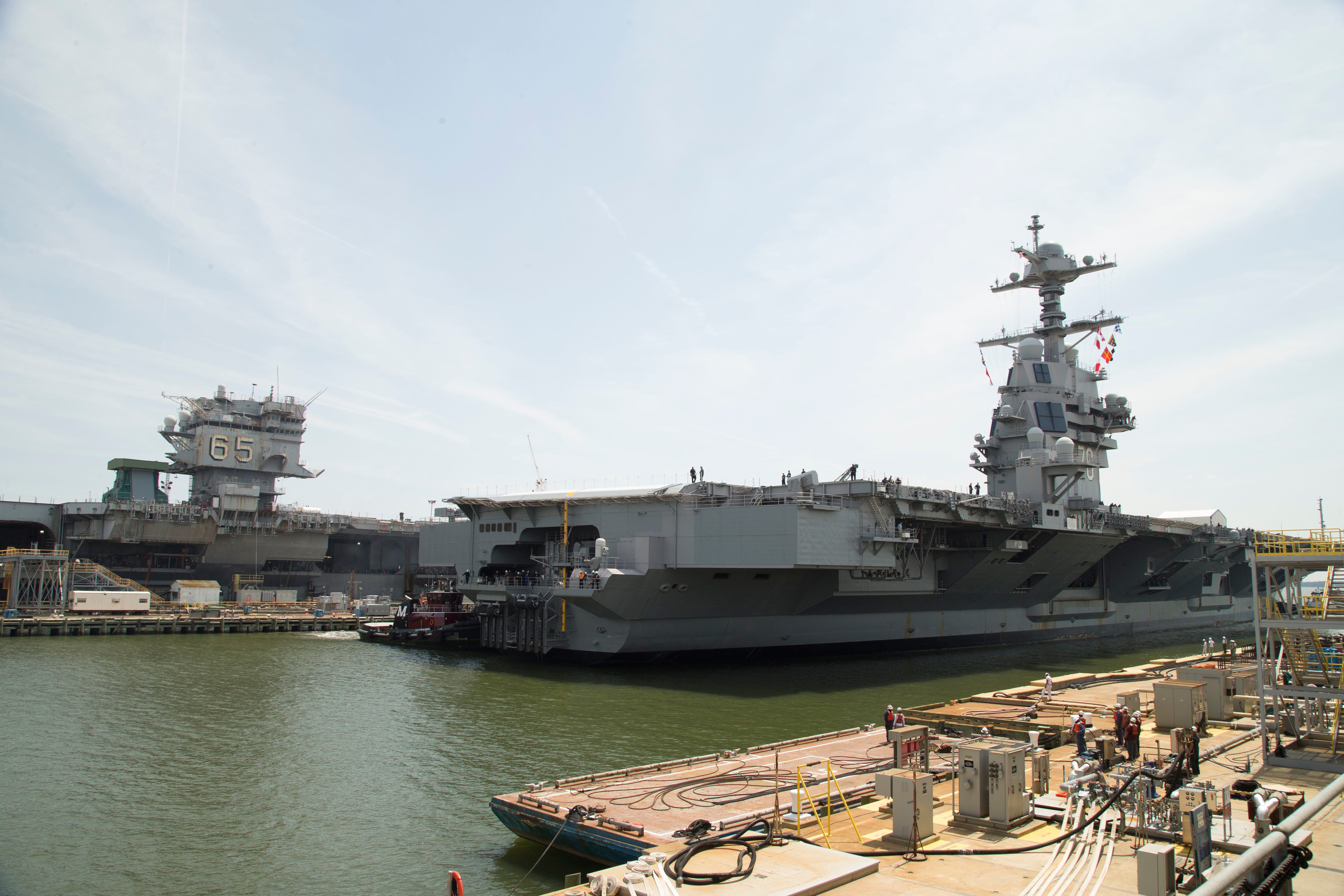 Two US Navy ships sit in water at a shipbuilding yard with a cloudy blue sky in the background.