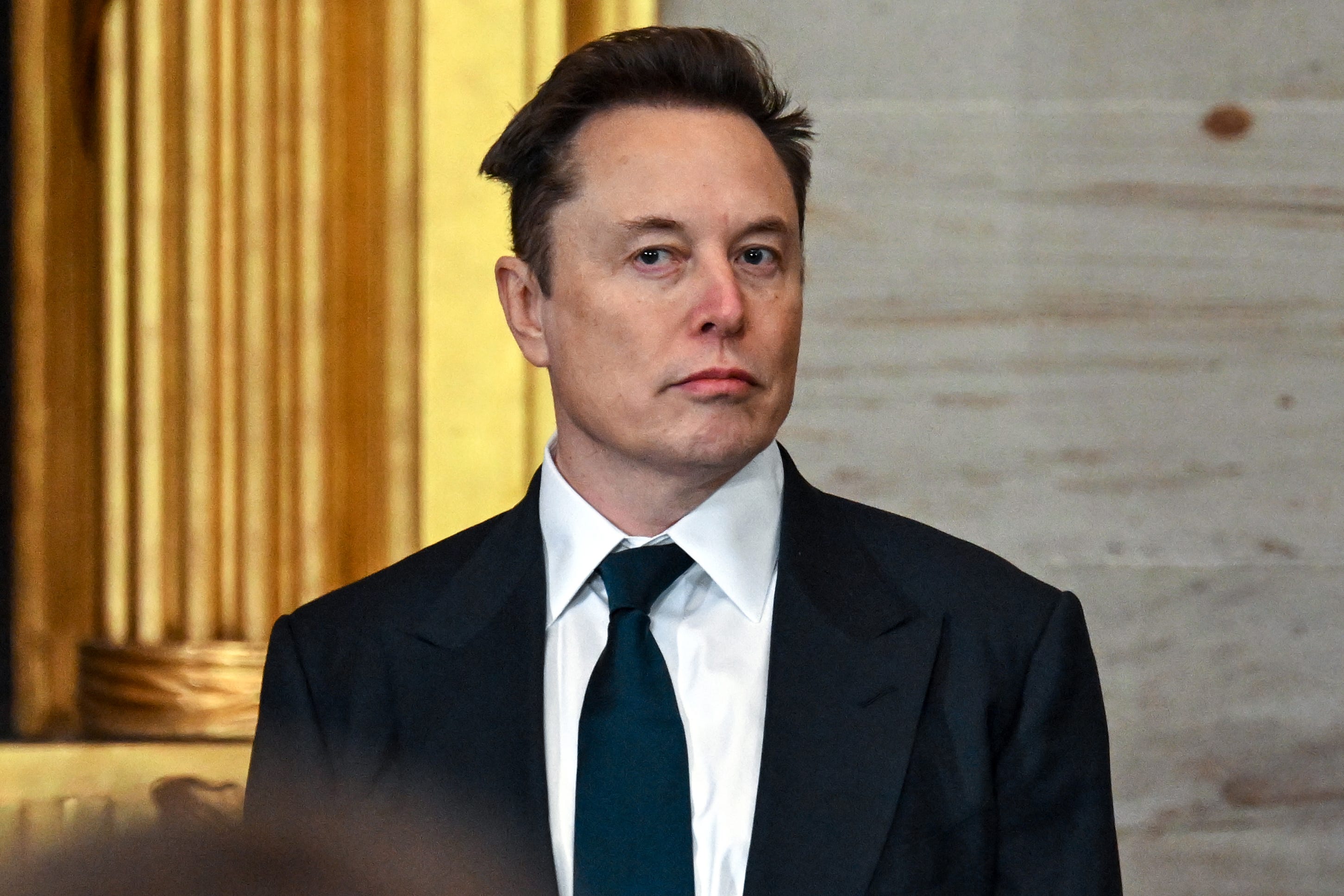 Elon Musk standing in a suit and tie with a straight face.