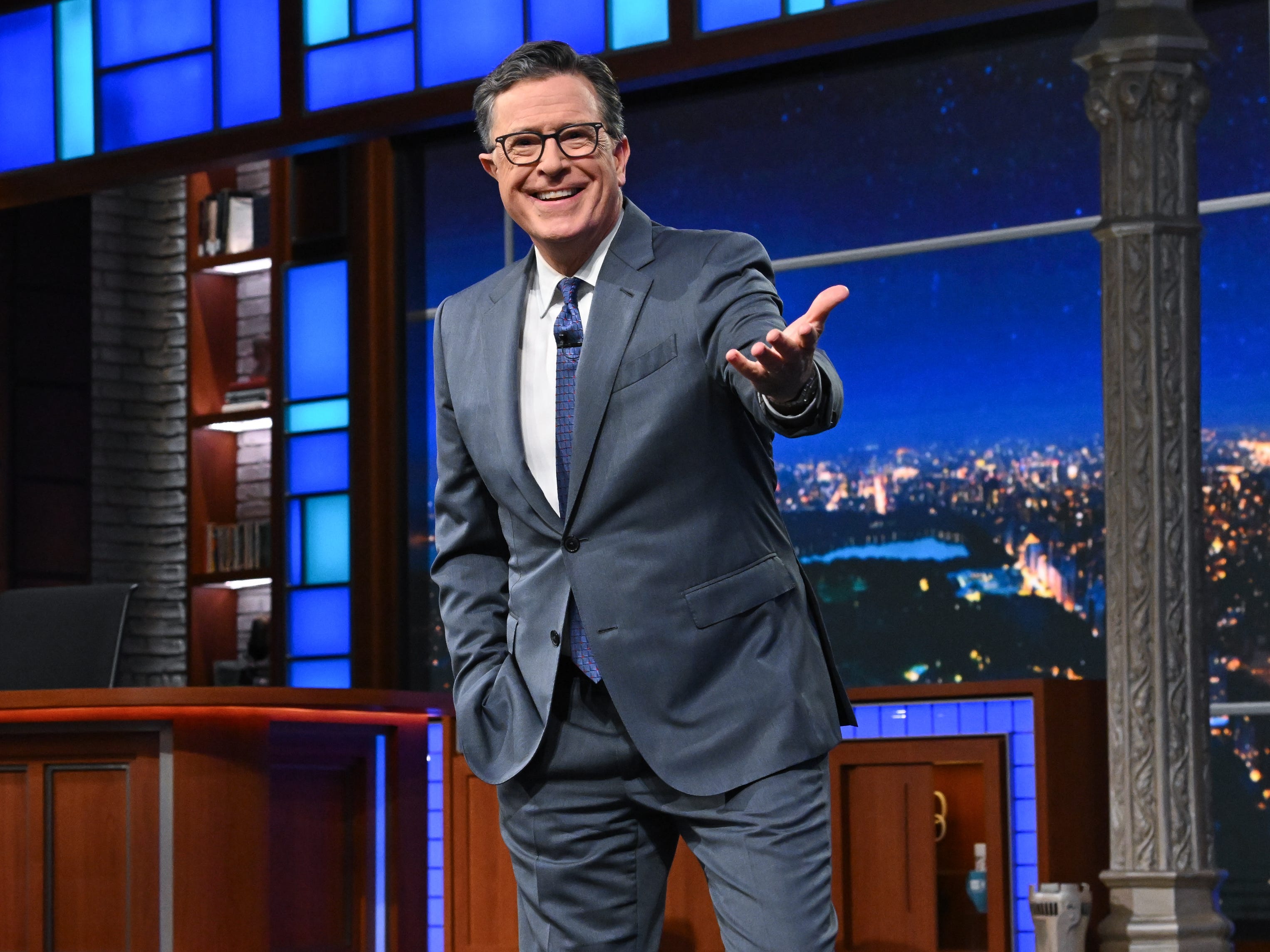 A middle-aged man with dark hair wearing a gray suit on a TV set. He's wearing black glasses and a metallic blue tie.