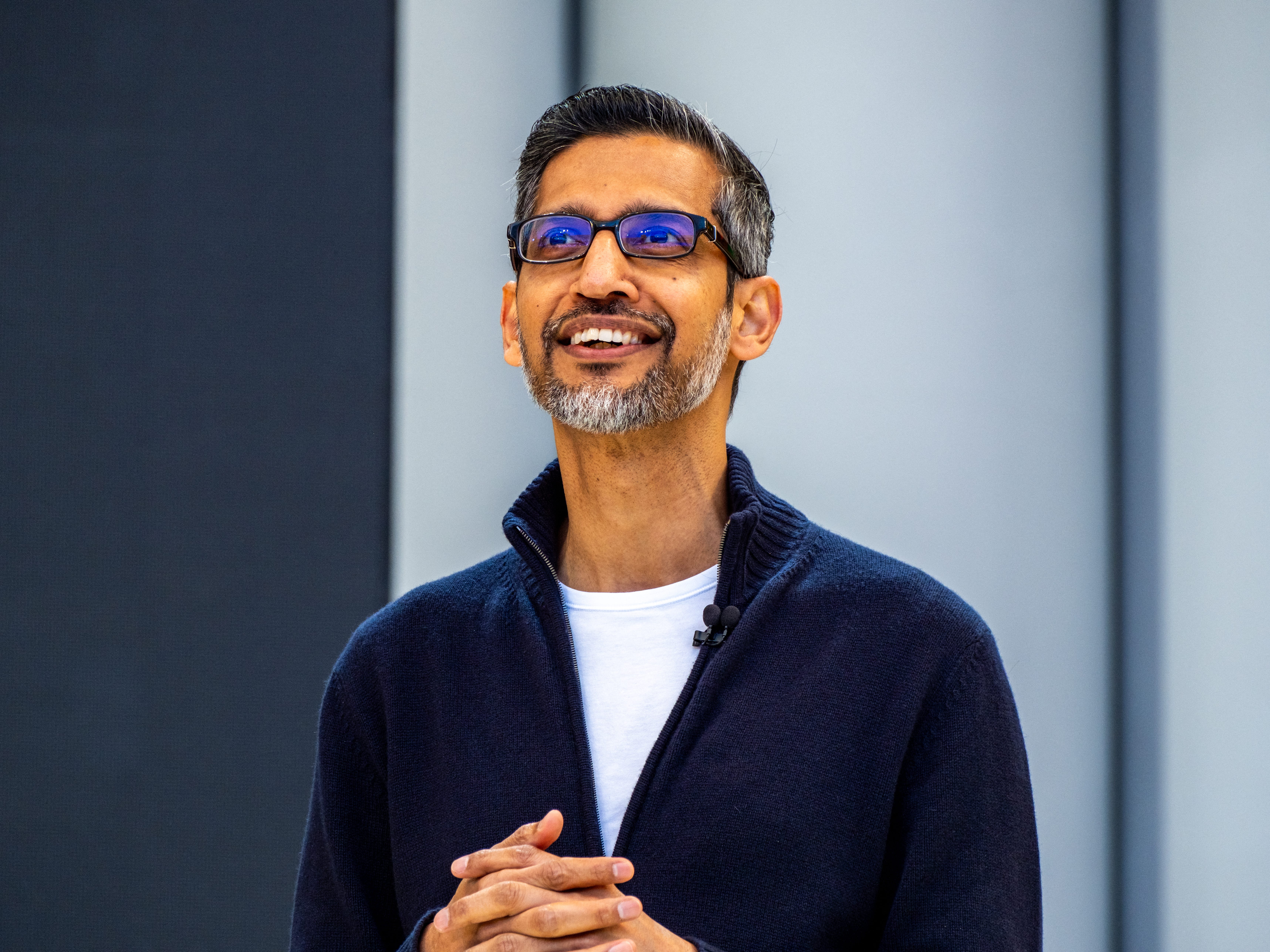 Google and Alphabet CEO Sundar Pichai looks upward, smiling, with his hands clasped in front of his stomach, wearing a blue zip sweater.