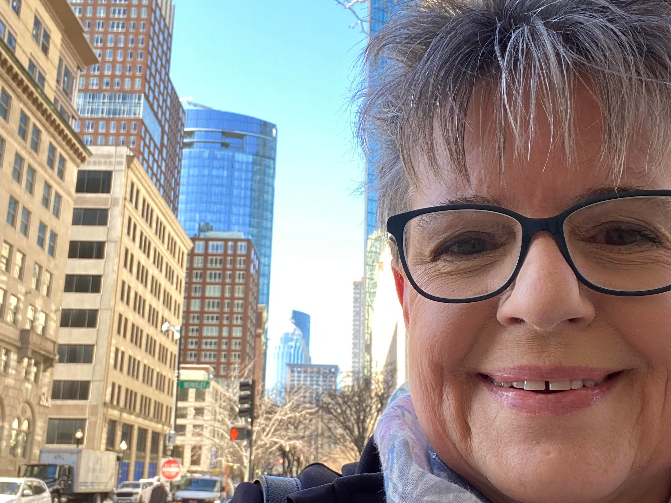 Woman with short grey hair and glasses takes a selfie in the street. Behind her are tall buildings, cars, and a clear blue sky.
