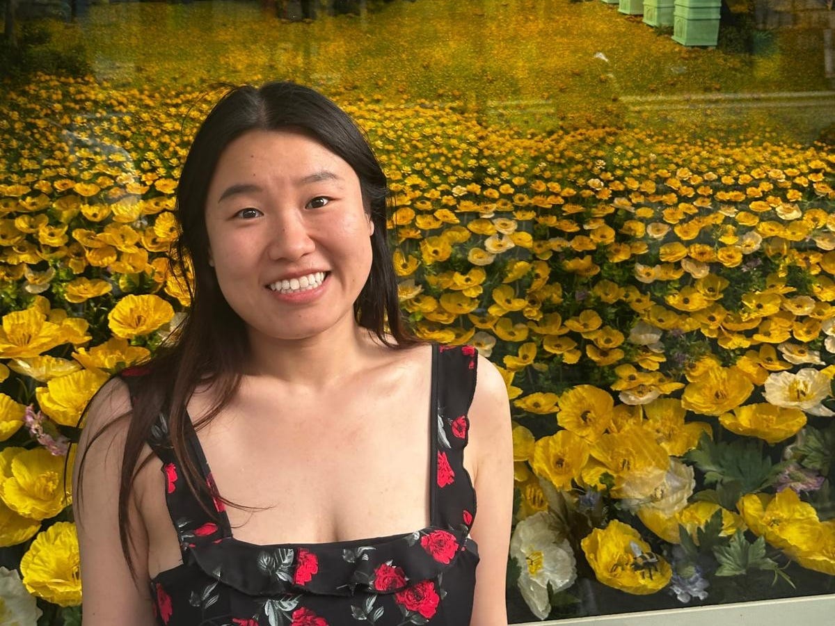 Kristine Wu smiling, standing in front of a yellow floral window, wearing a black dress with red flowers.