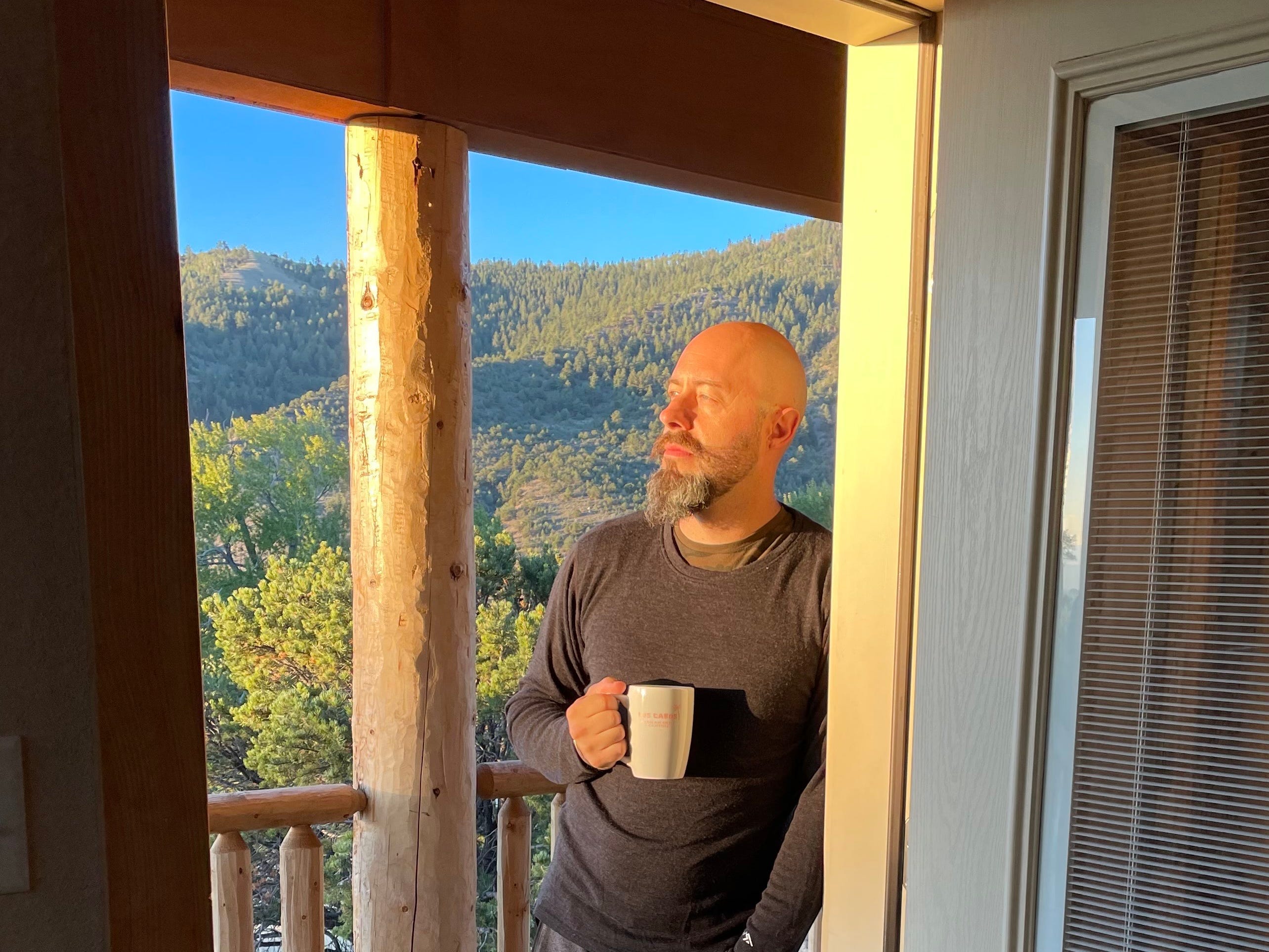 a man on a porch holding a mug and looking off to the left. In the background is a scenic mountain view