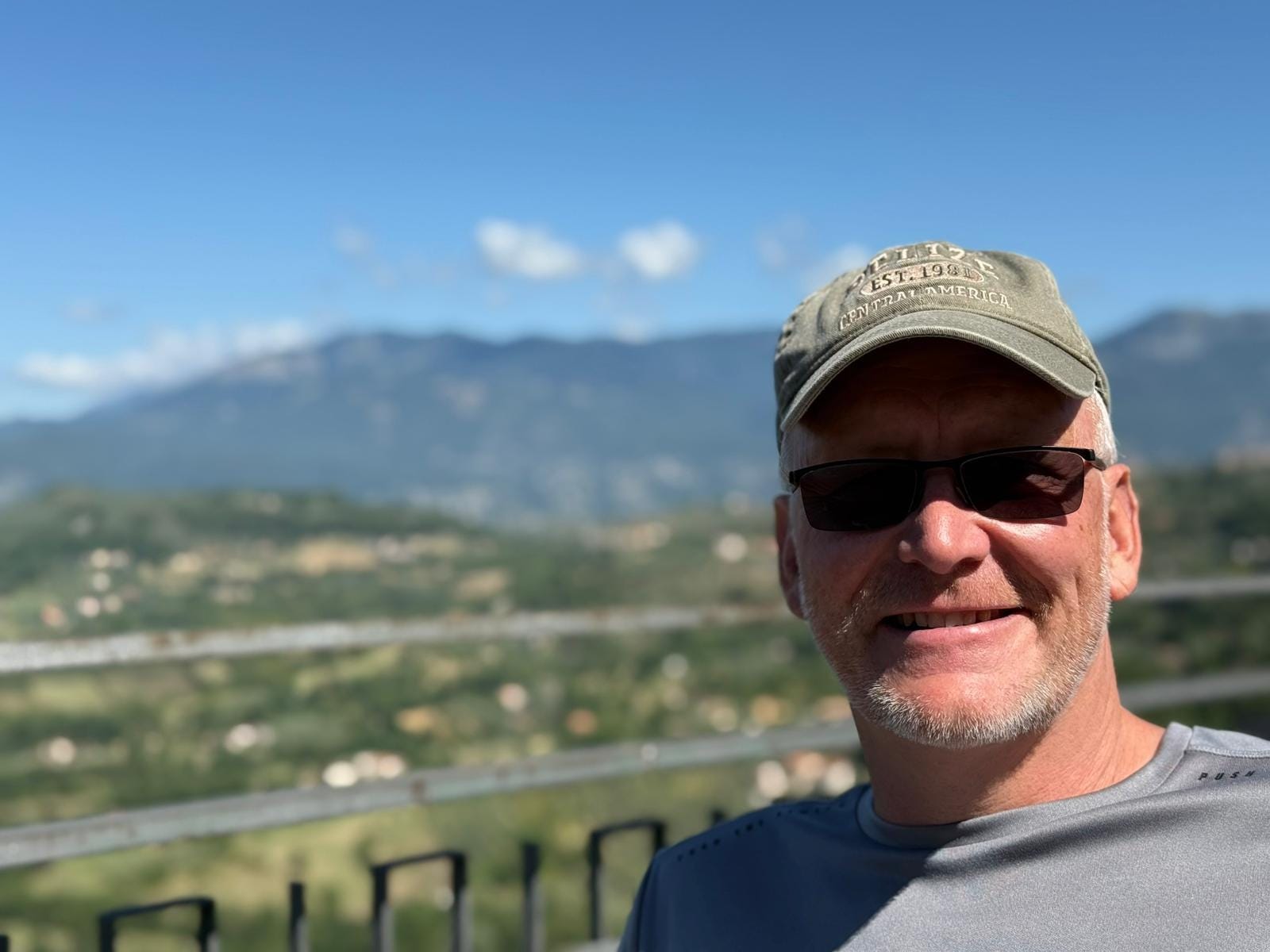 A man wearing a baseball hat smiles in front of rolling hills.