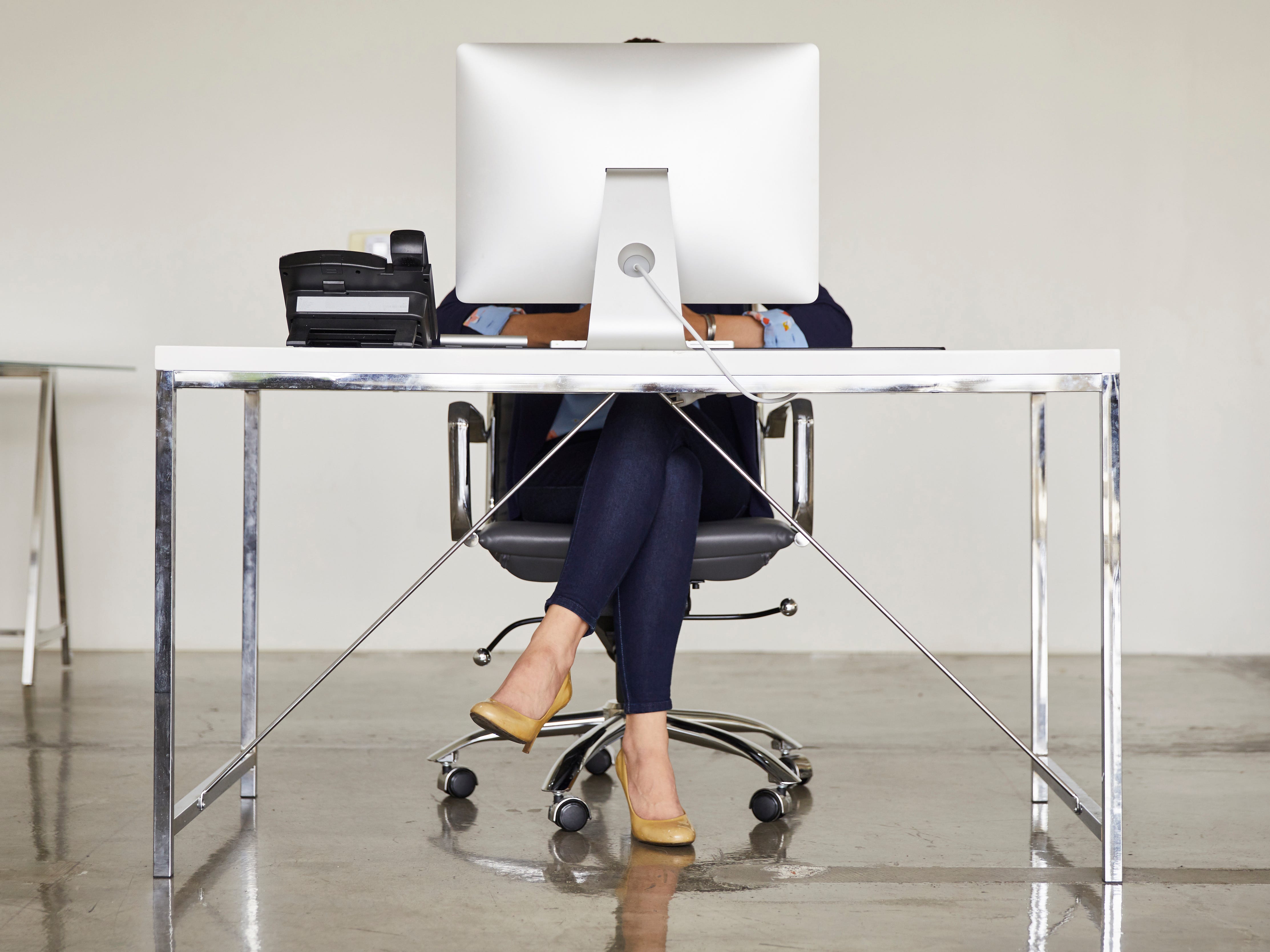 A woman wearing blue pants and tan heels sits at a modern desk with a computer and a phone obscuring her face.