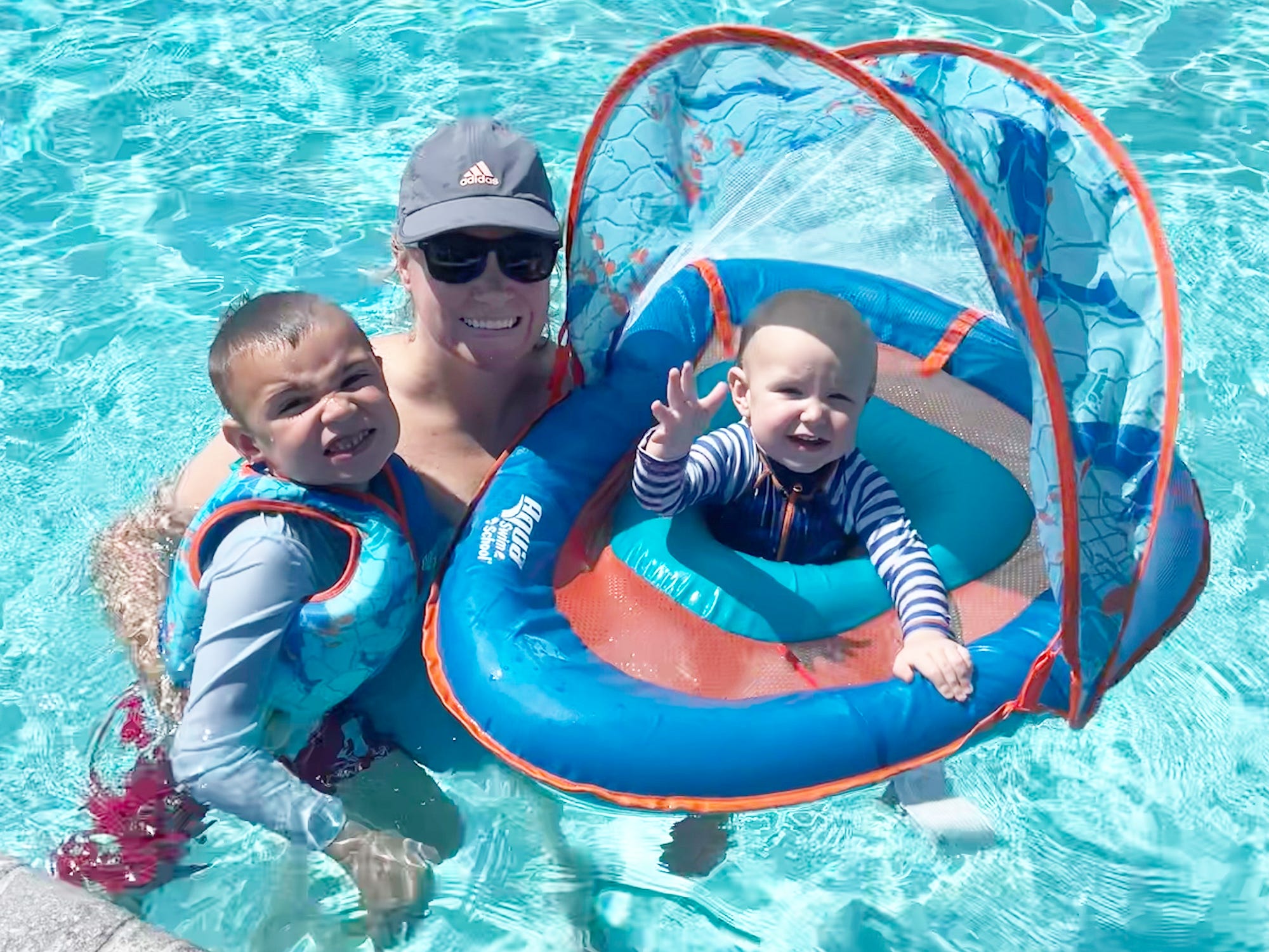 A mother swims in a pool with two young kids. One of them in in a baby float.