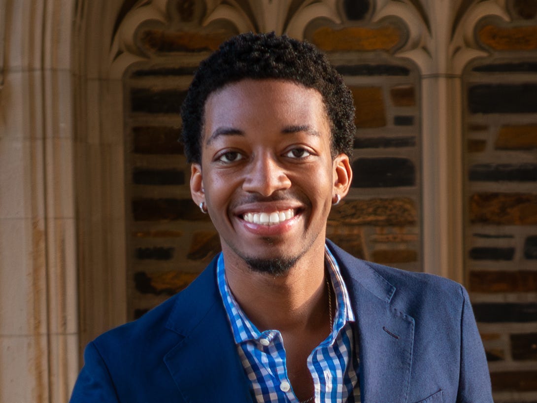 Photo of a man with black curly hair wearing a blue blazer and checkered collared button-down smiling for a photo.