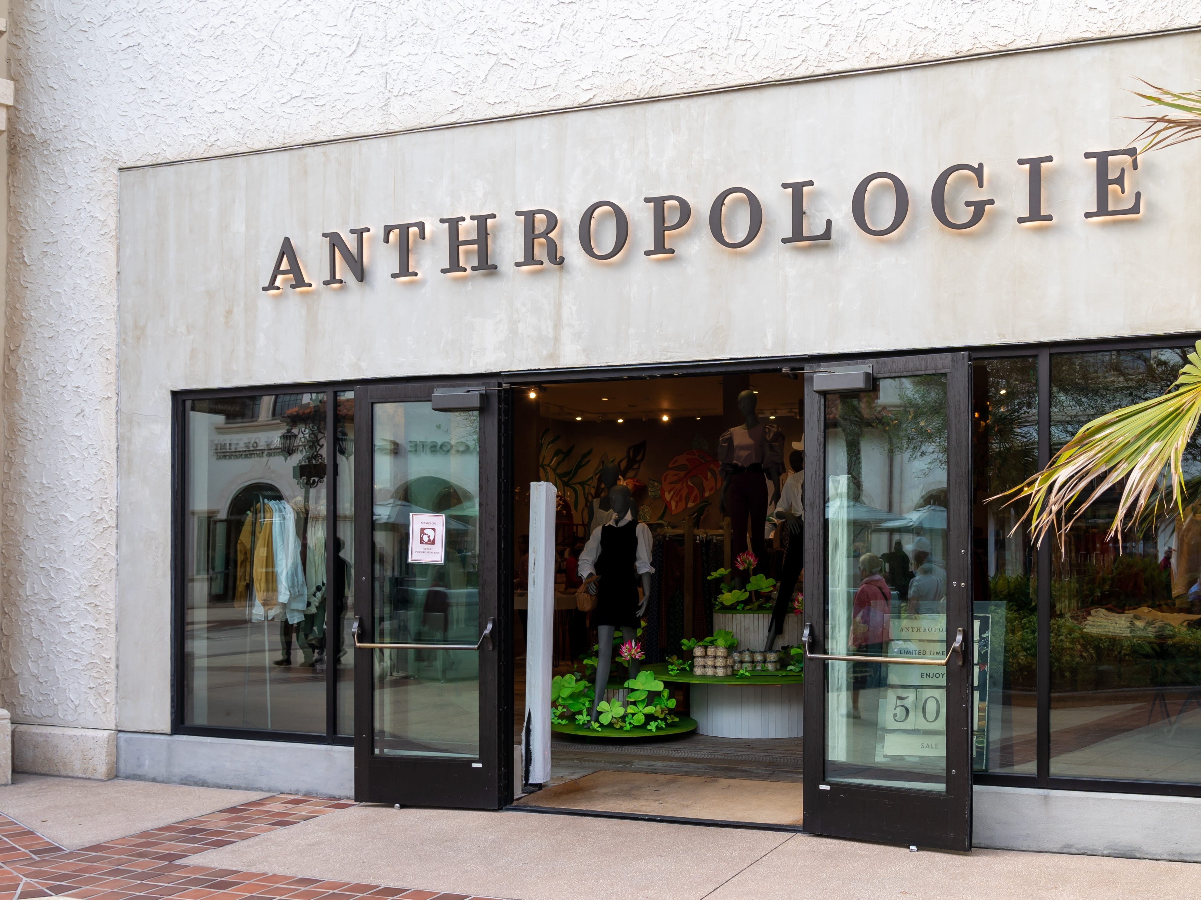 Gray building exterior of Anthropologie store. A palm tree and clothing with plant decor inside the store is visible