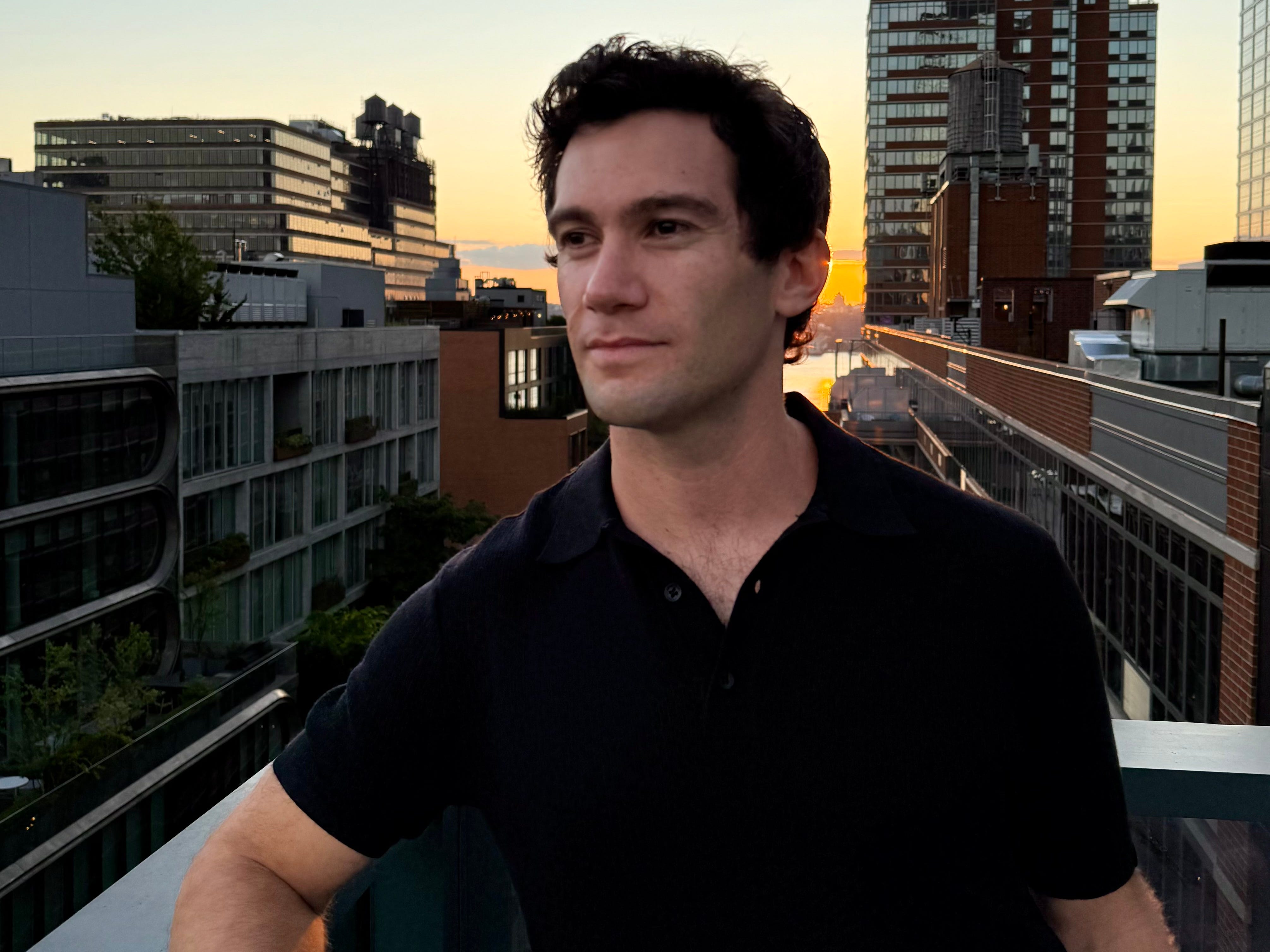 A man with dark hair and a short sleeve polo standing on a rooftop looking off to the side. A sunset skyline with buildings is in the background