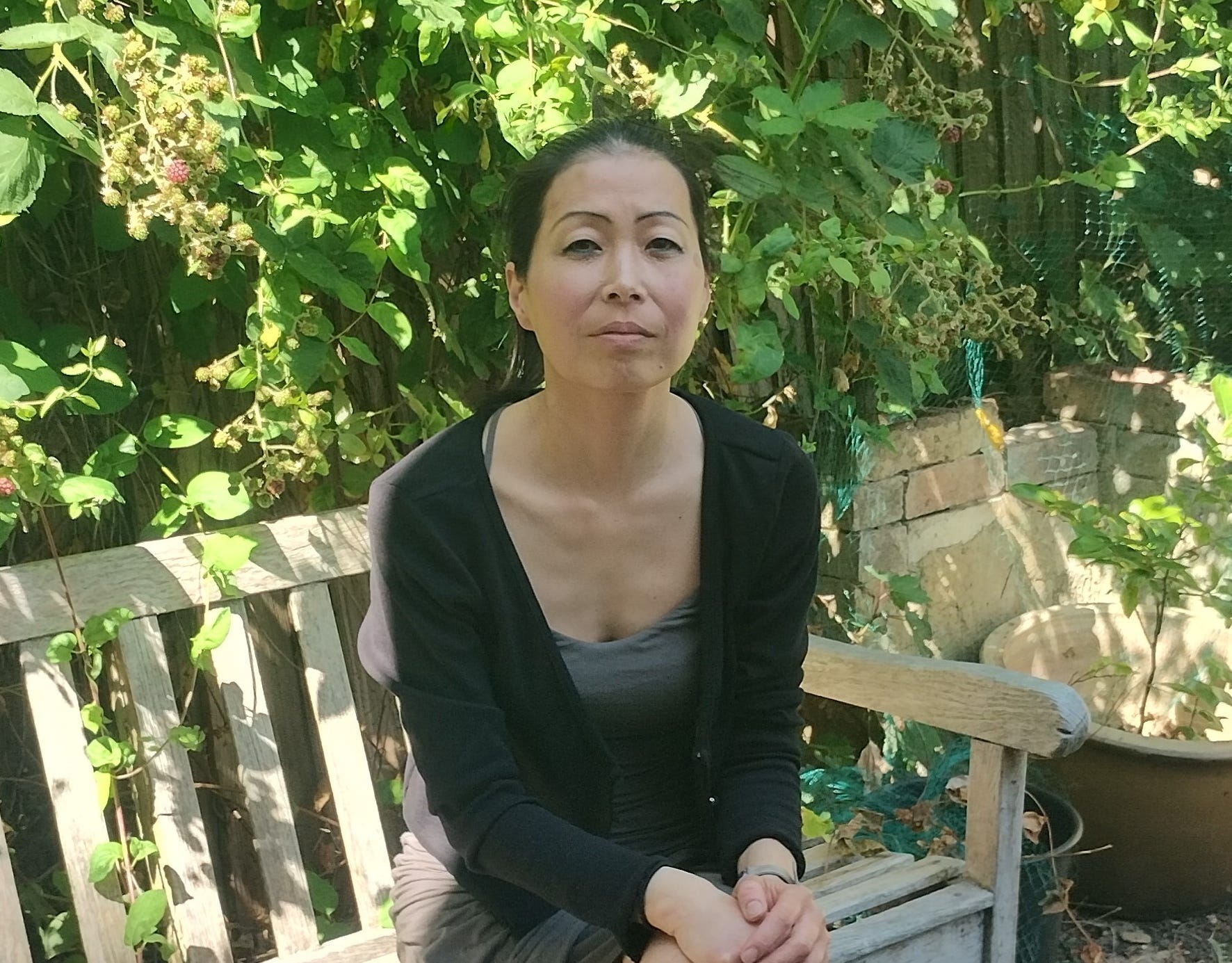 Risa Morimoto sitting on a bench outdoors in front of some greenery.