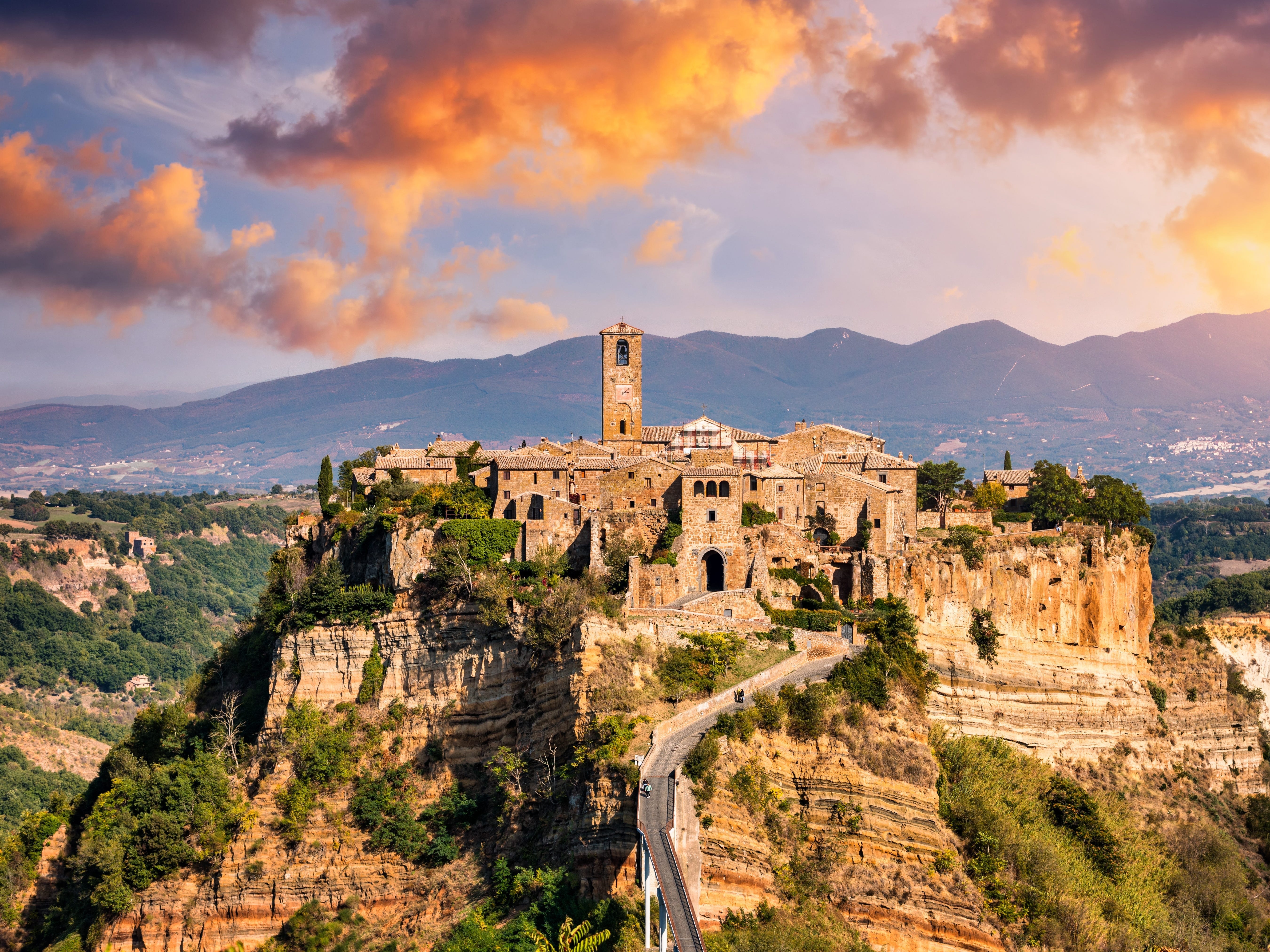 A medieval Italian city on a hill at sunset, with mountains in the background.