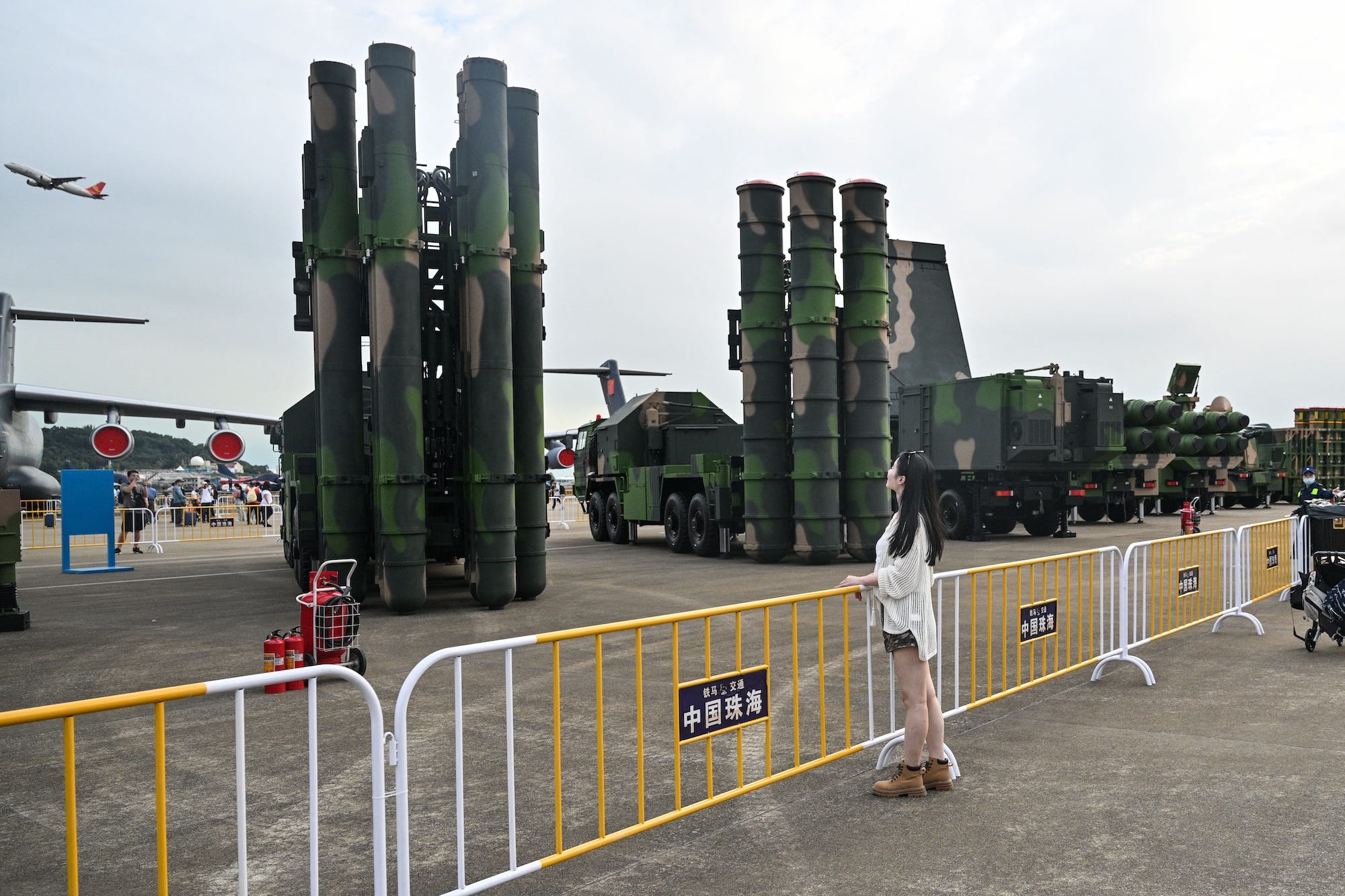 A woman stands in front of a yellow and white metal barrier in front of two large pieces of weapnry made up of large cylinders pointed towards the sky