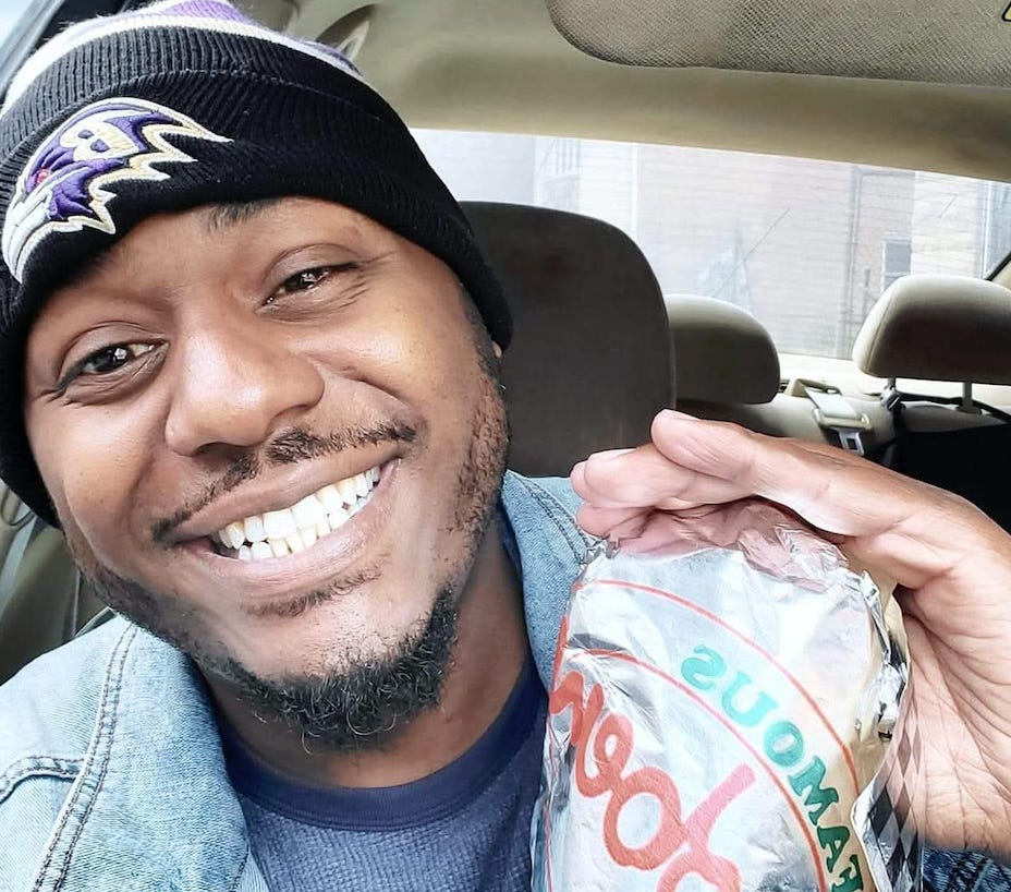 Kreskin Torres smiles while he wears a Baltmore Ravens beanie and denim jacket as he smiles and holds a wrapped sandwich while sitting in a car.