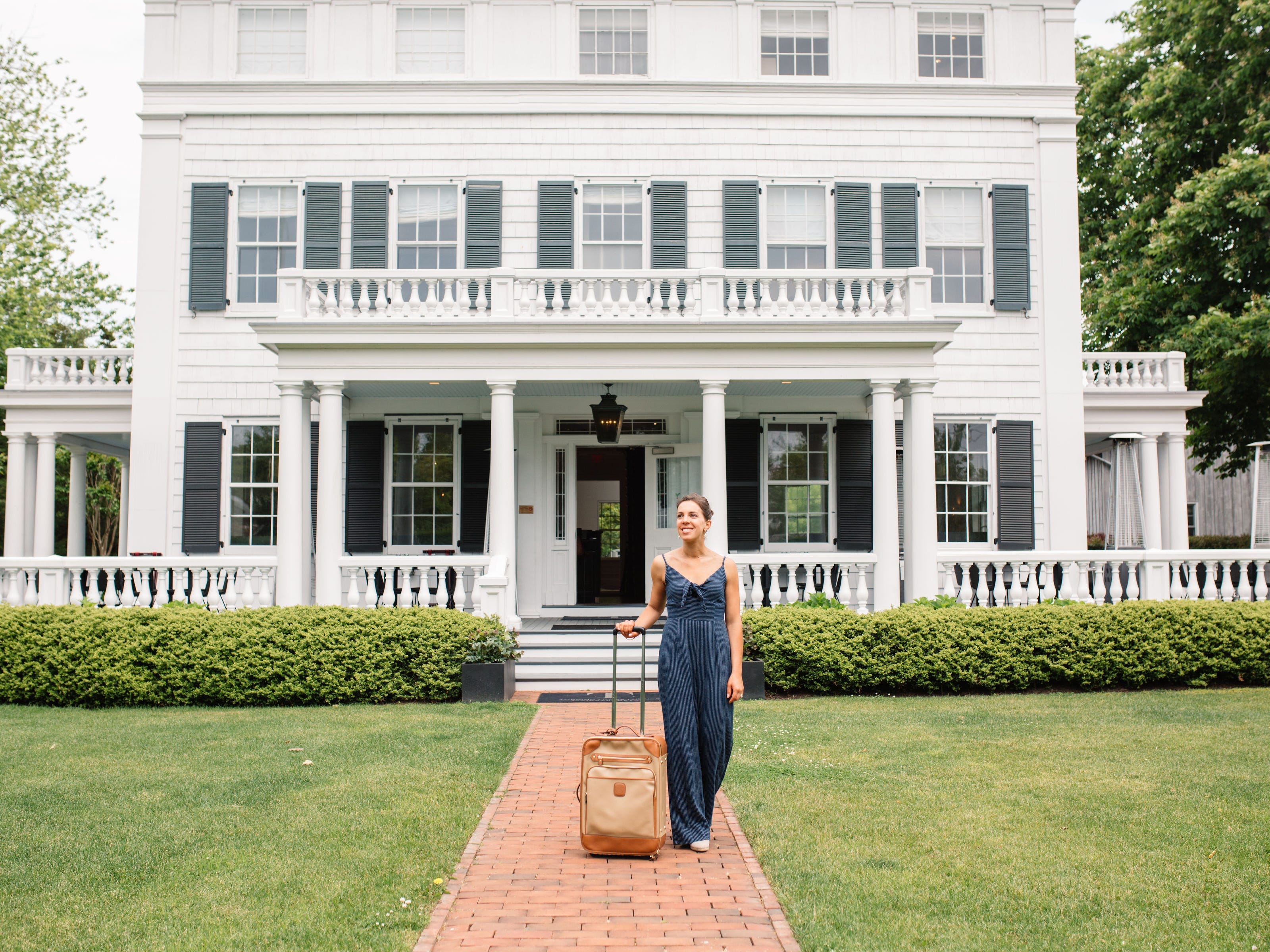 Vanessa stands in front of a large white house with a suitcase.