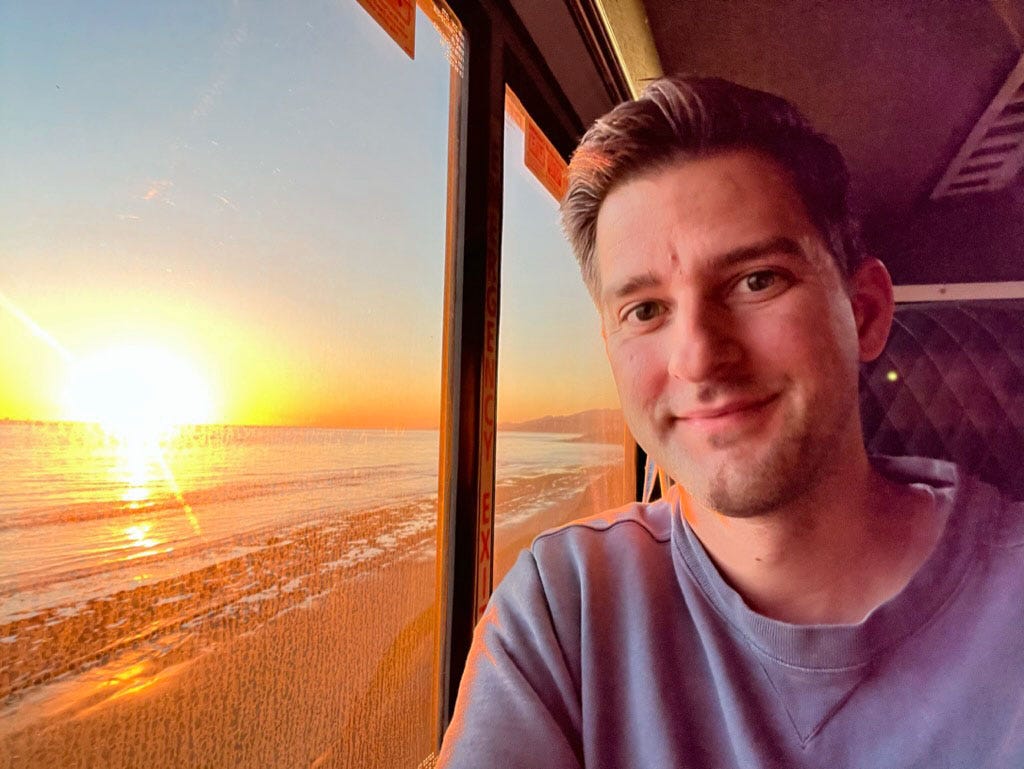 William takes a selfie near a train window with a view of the beach at sunset.