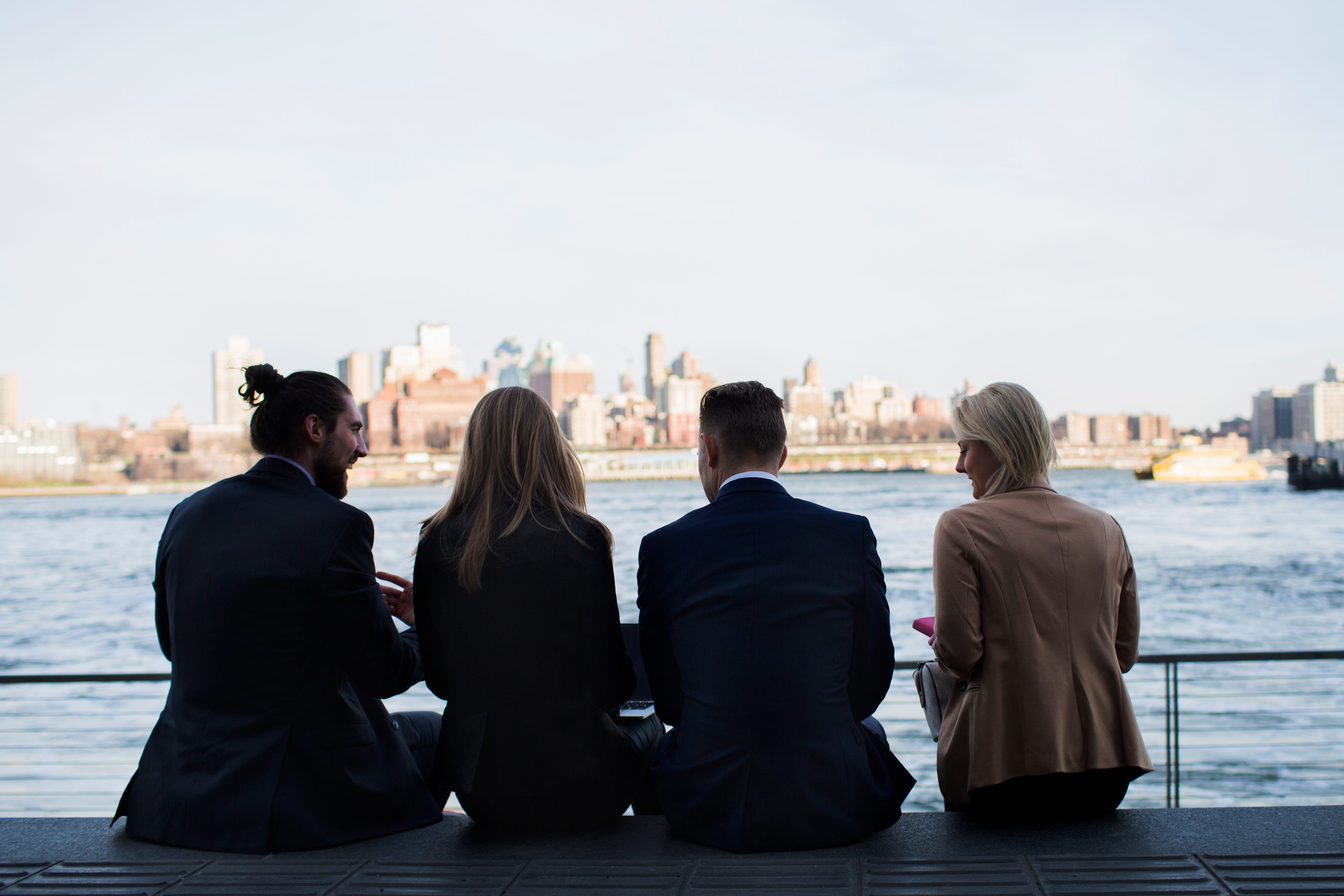 four people sit with their back to the camera overlooking a river and city skyline